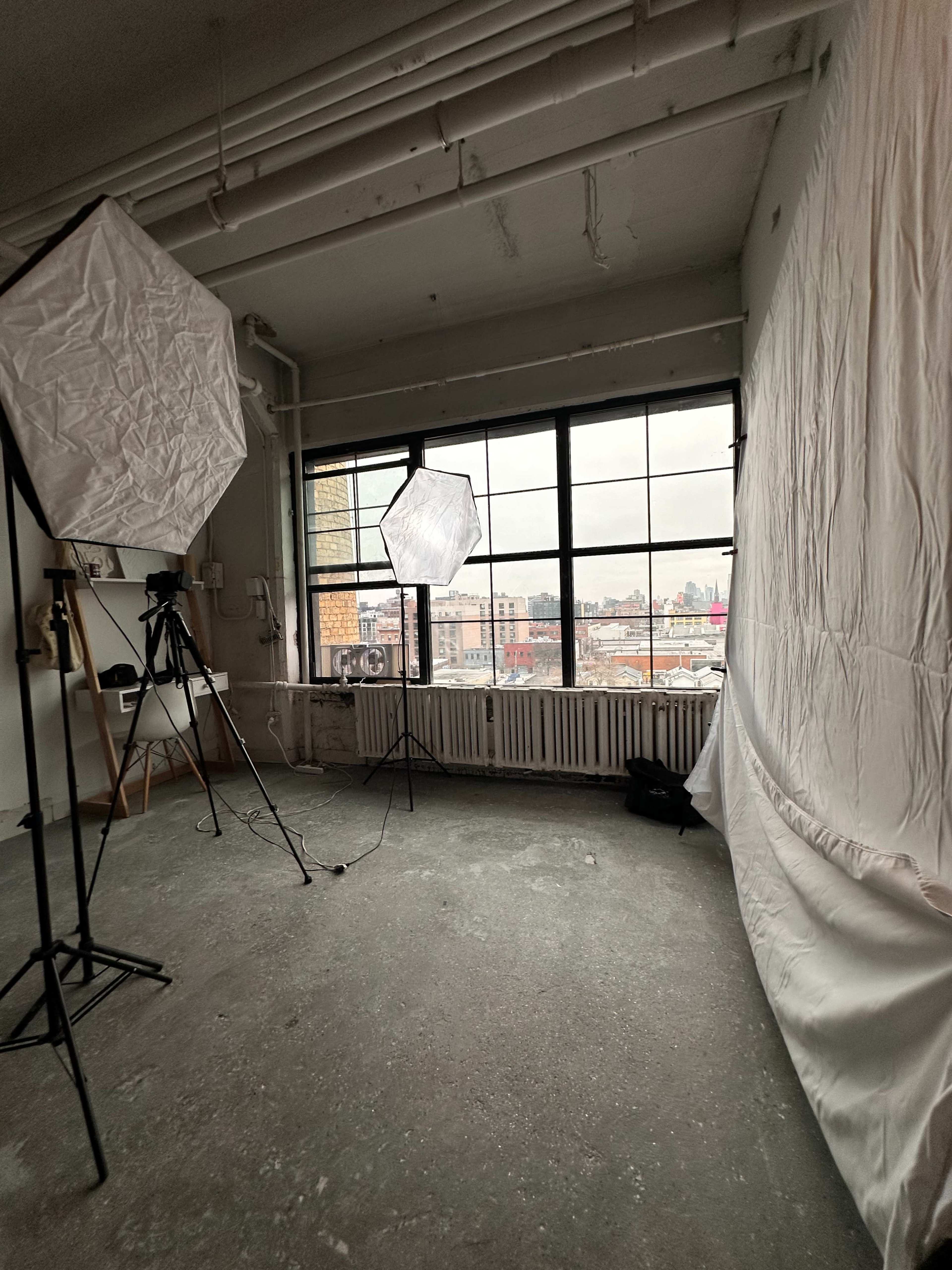 The image shows a photography studio with two softbox lights, a camera on a tripod, and a white backdrop near a large window overlooking an urban landscape.