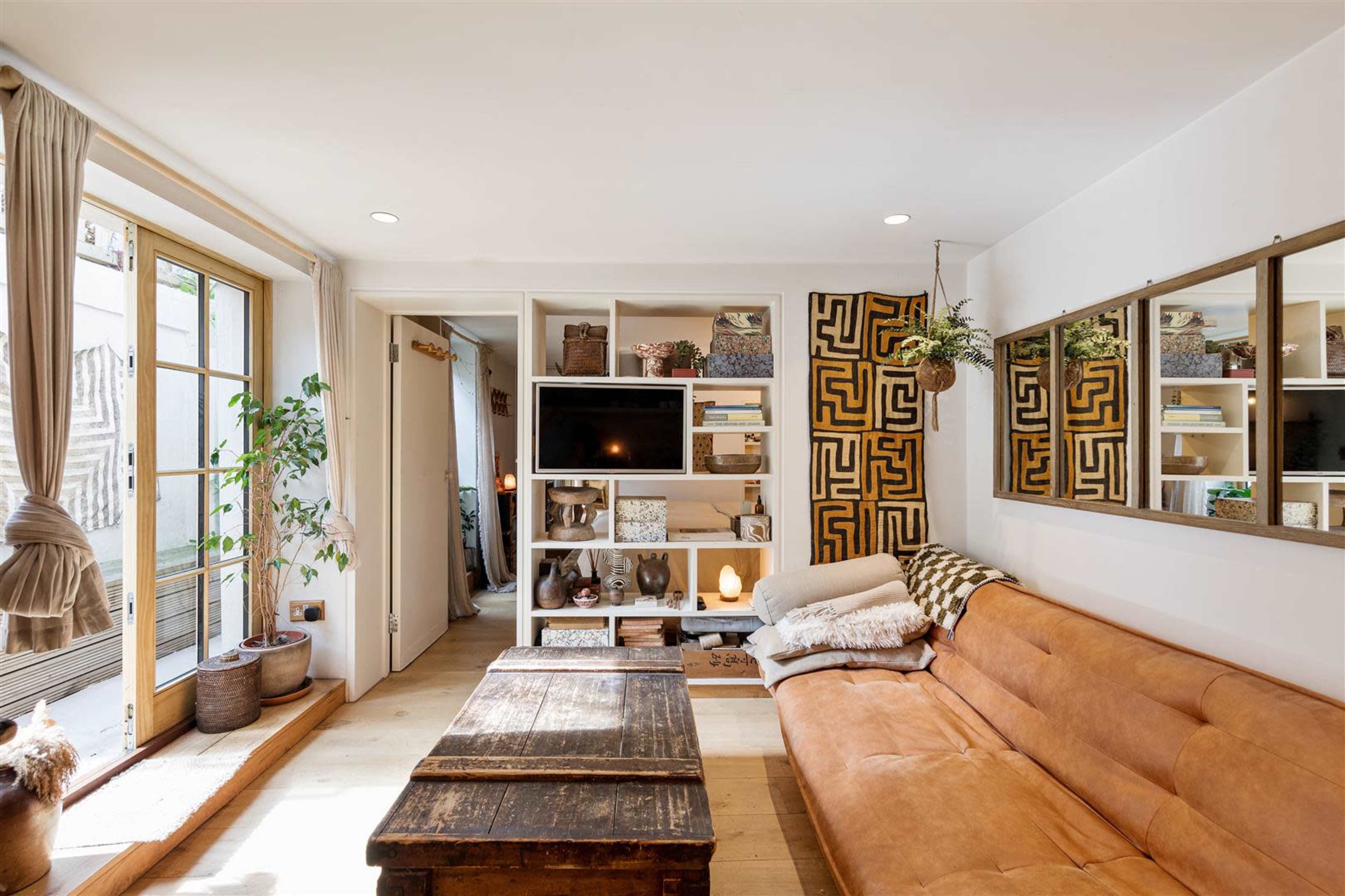 A cozy living room features a brown couch, wooden coffee table, wall-mounted television, and shelves filled with decorative items and plants.