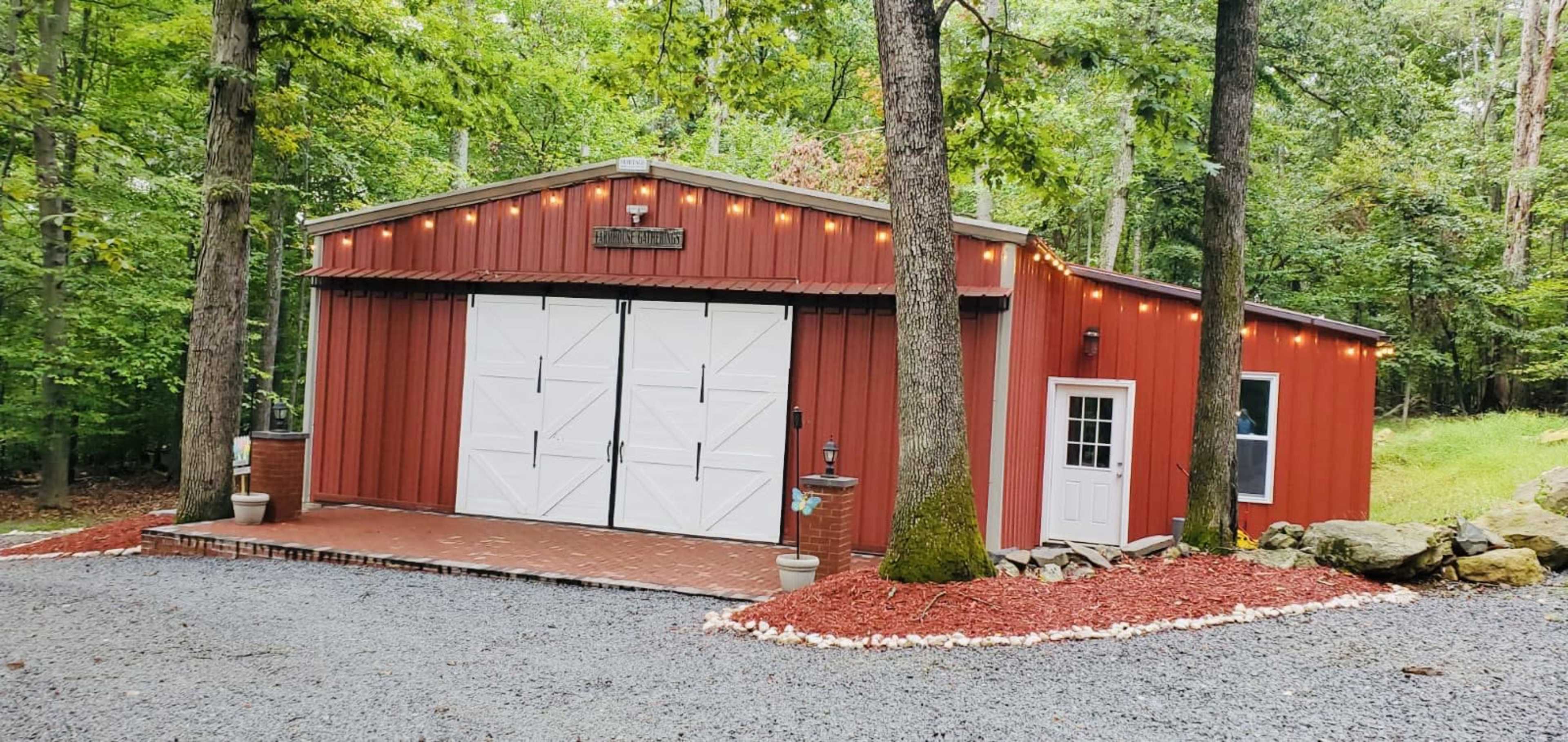 A red metal garage with white double doors and string lights is surrounded by greenery and gravel landscaping.
