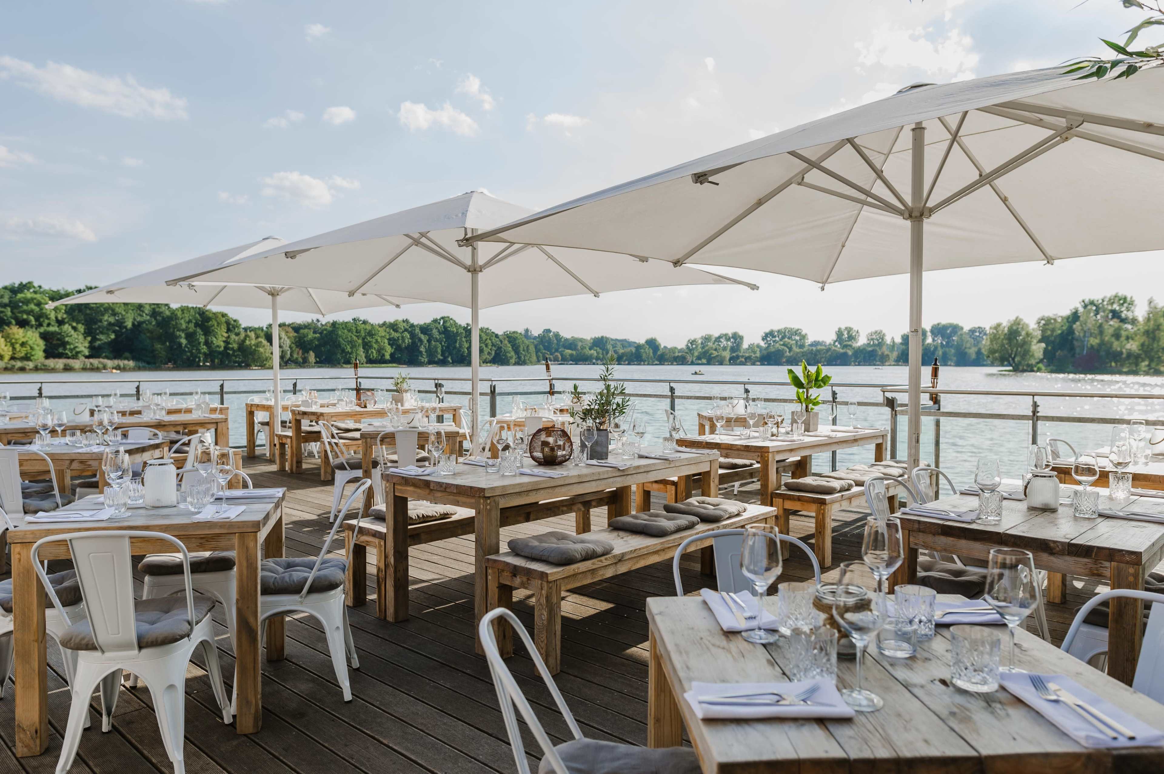 An outdoor dining area with wooden tables and chairs, shaded by large umbrellas, overlooks a body of water surrounded by greenery.