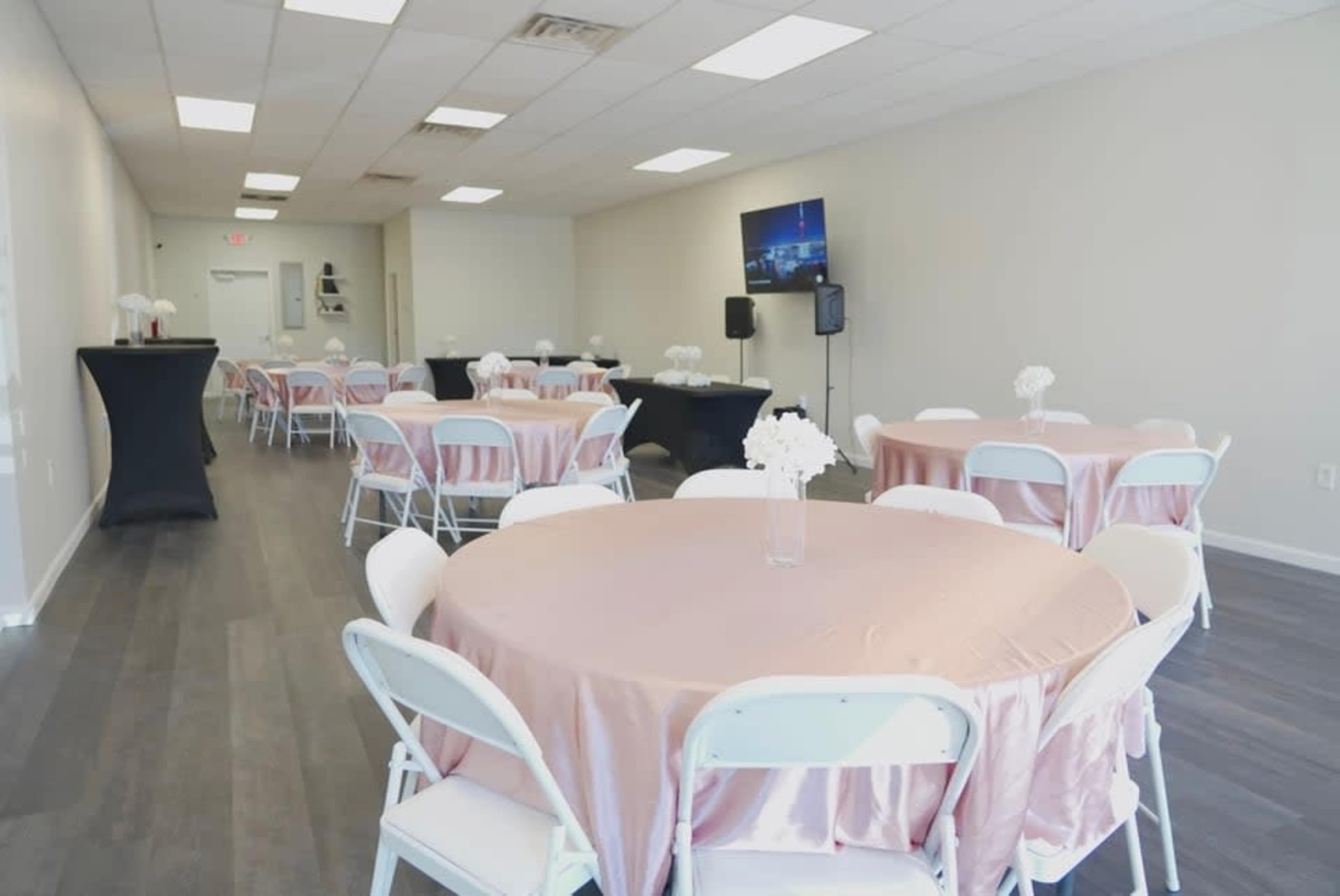 The image shows a spacious event room with round tables covered in pink tablecloths and white folding chairs arranged neatly around them.