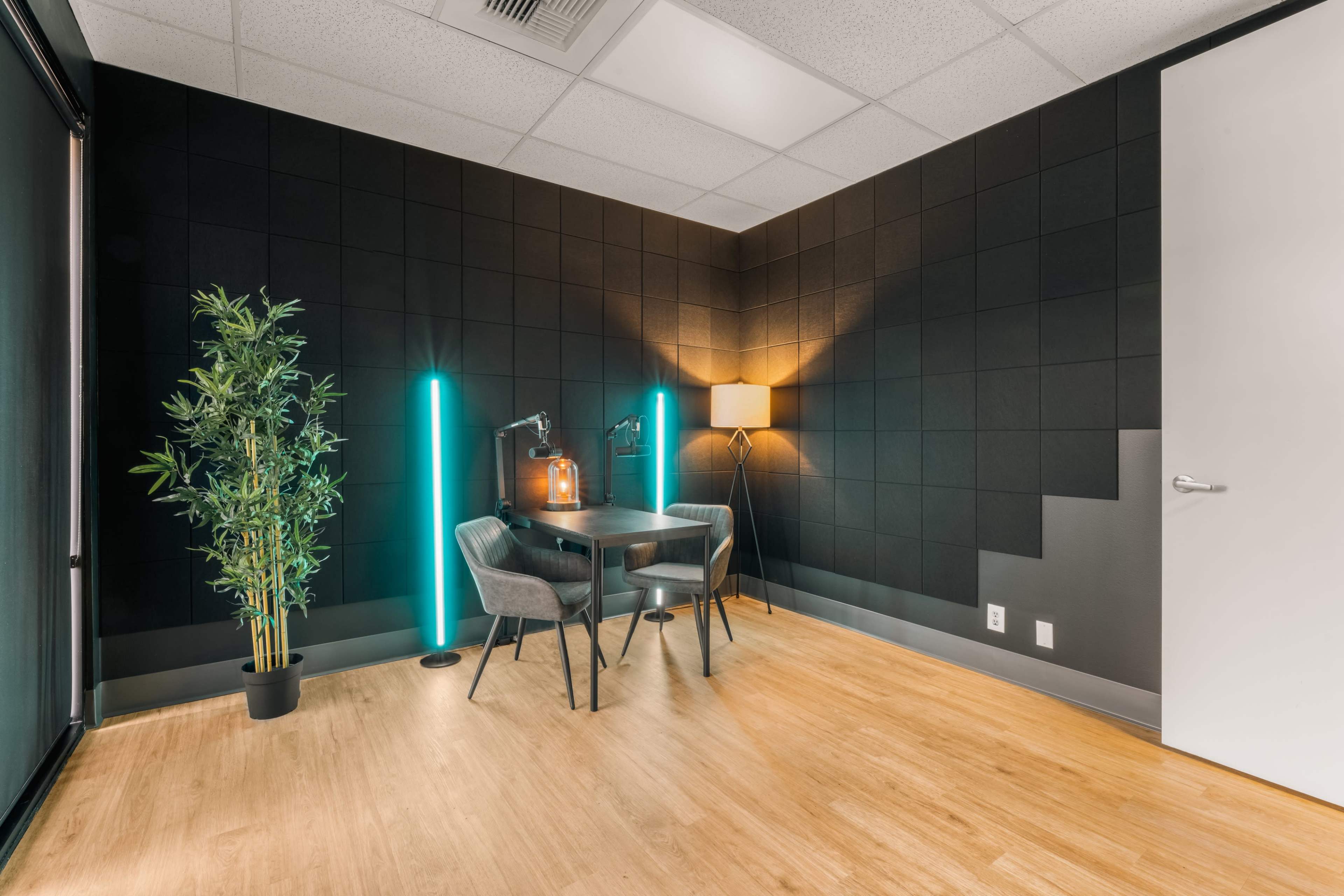 A small office space features a black wall, a wooden floor, a desk with two chairs, and lighting from a lamp and two standing lights.