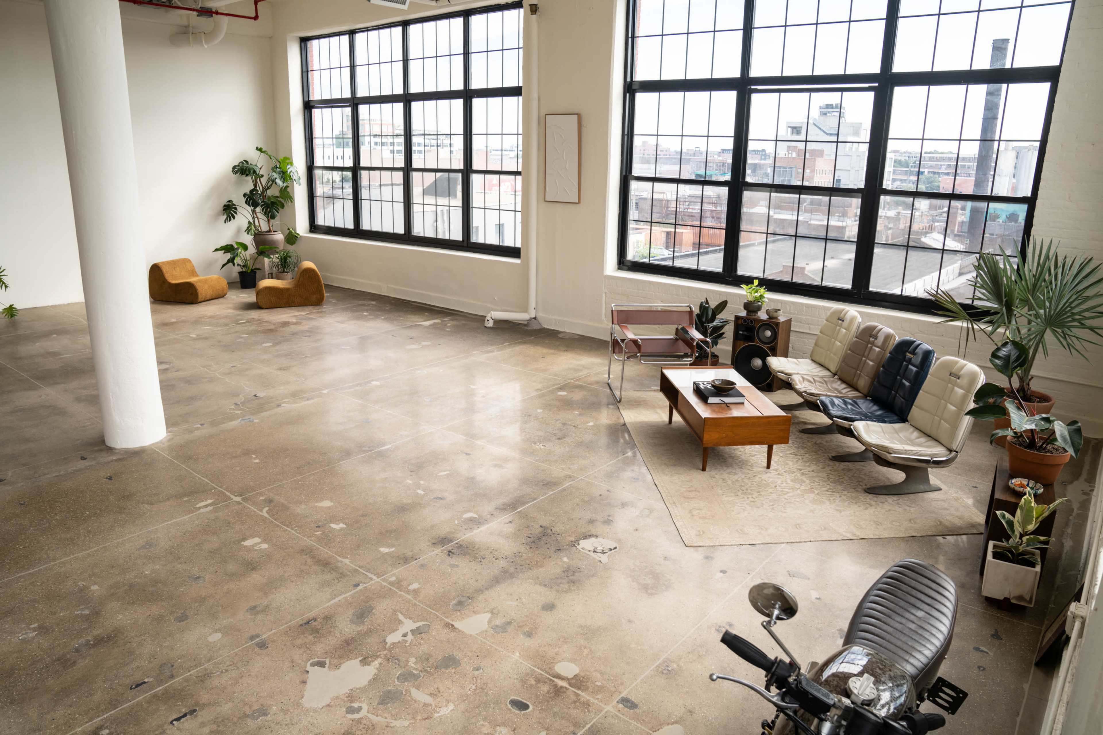 The image shows a spacious, industrial-style room with large windows, featuring furniture such as a leather sofa, a wooden coffee table, and a motorcycle in the corner.