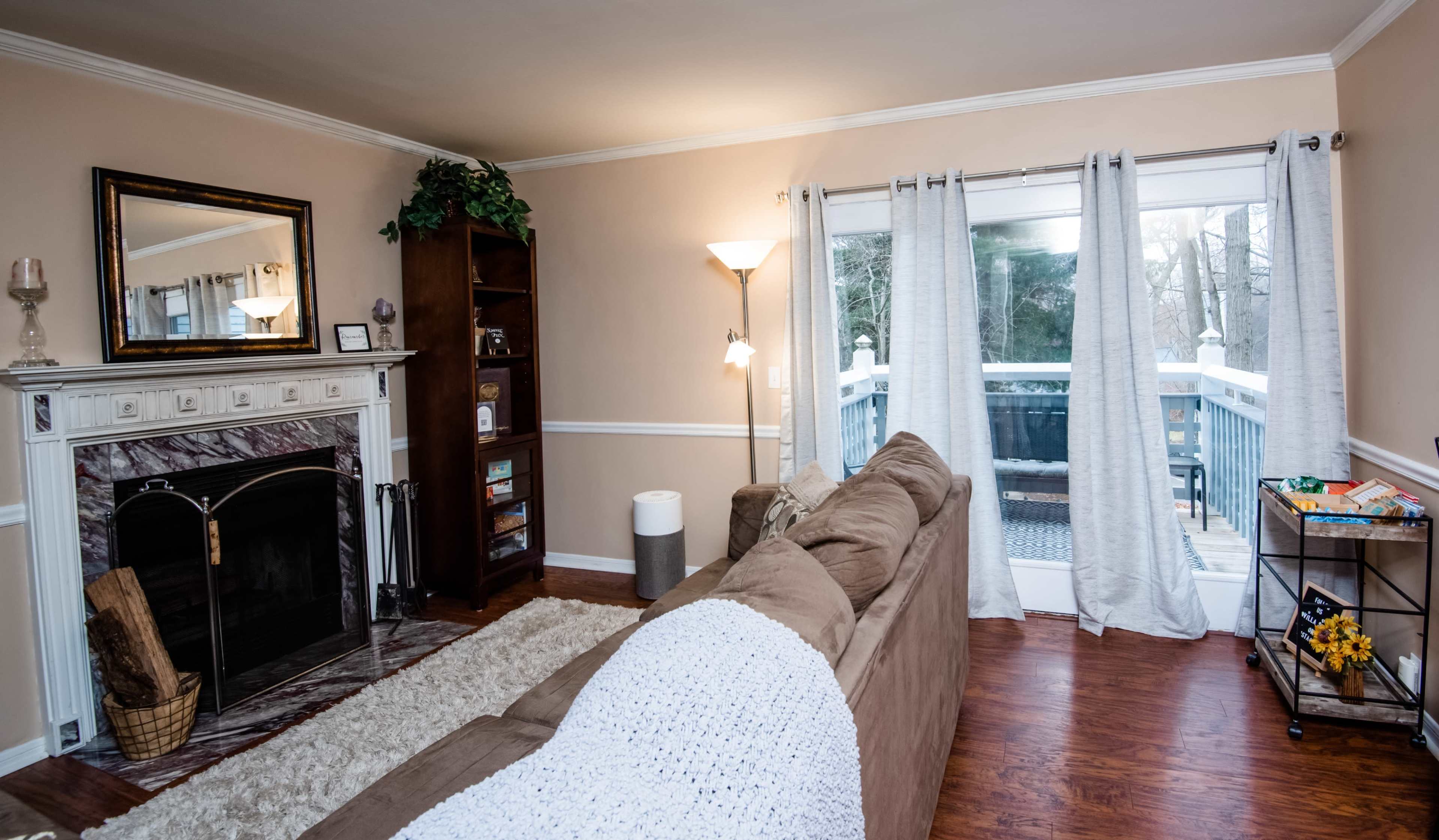 The living room features a brown sofa facing a fireplace, with a mirror above it, a bookshelf, and a sliding glass door leading to a deck.