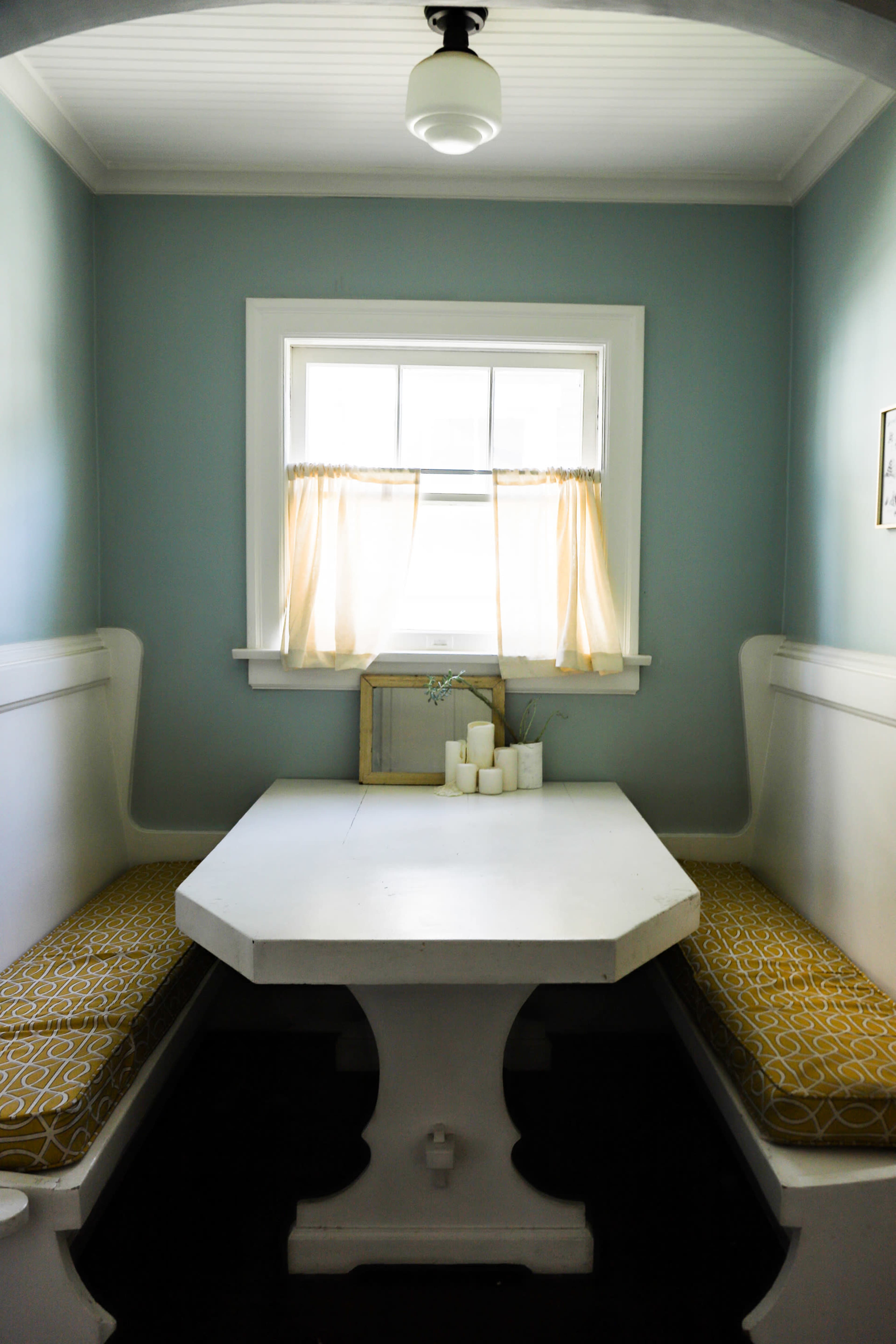 The image shows a small dining nook with an octagonal white table and two built-in benches with patterned cushions, illuminated by natural light from a nearby window.