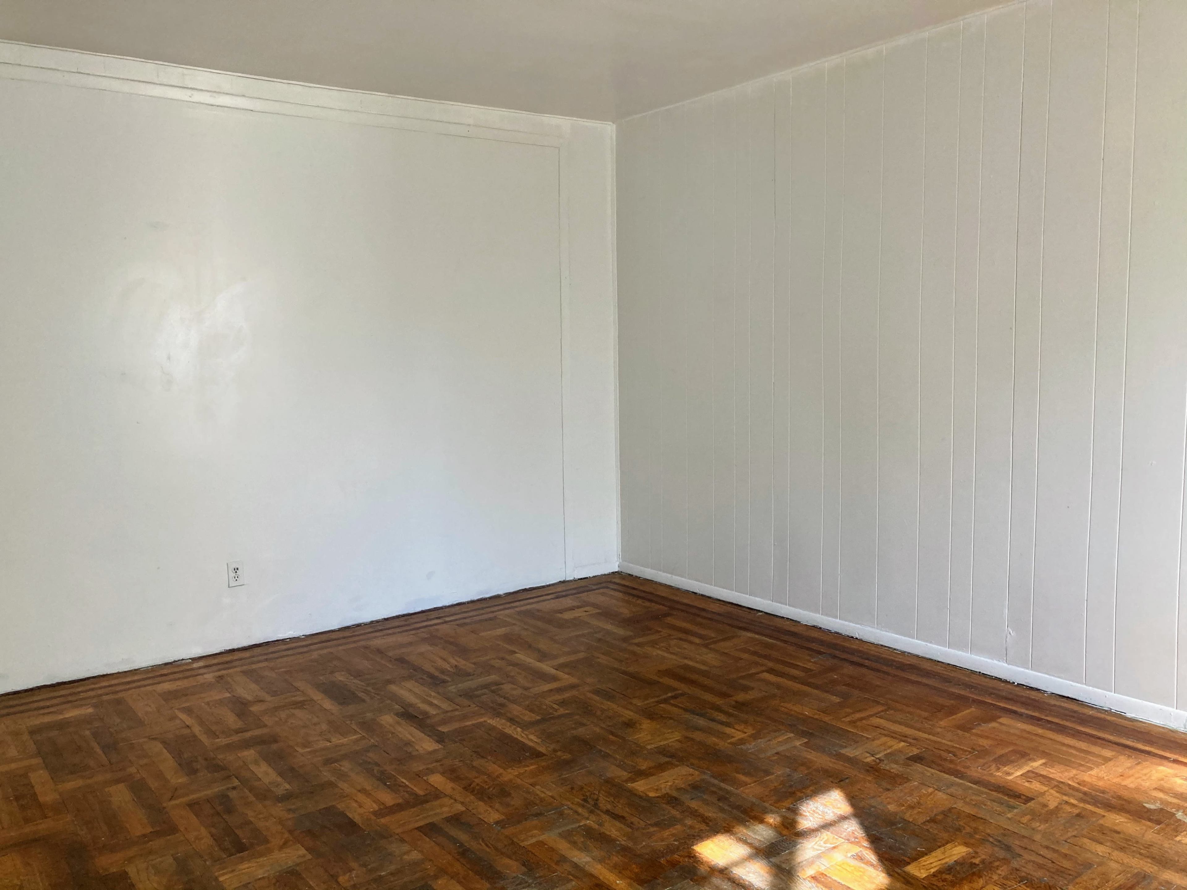 The image shows a bare room with wooden parquet flooring and two white walls.