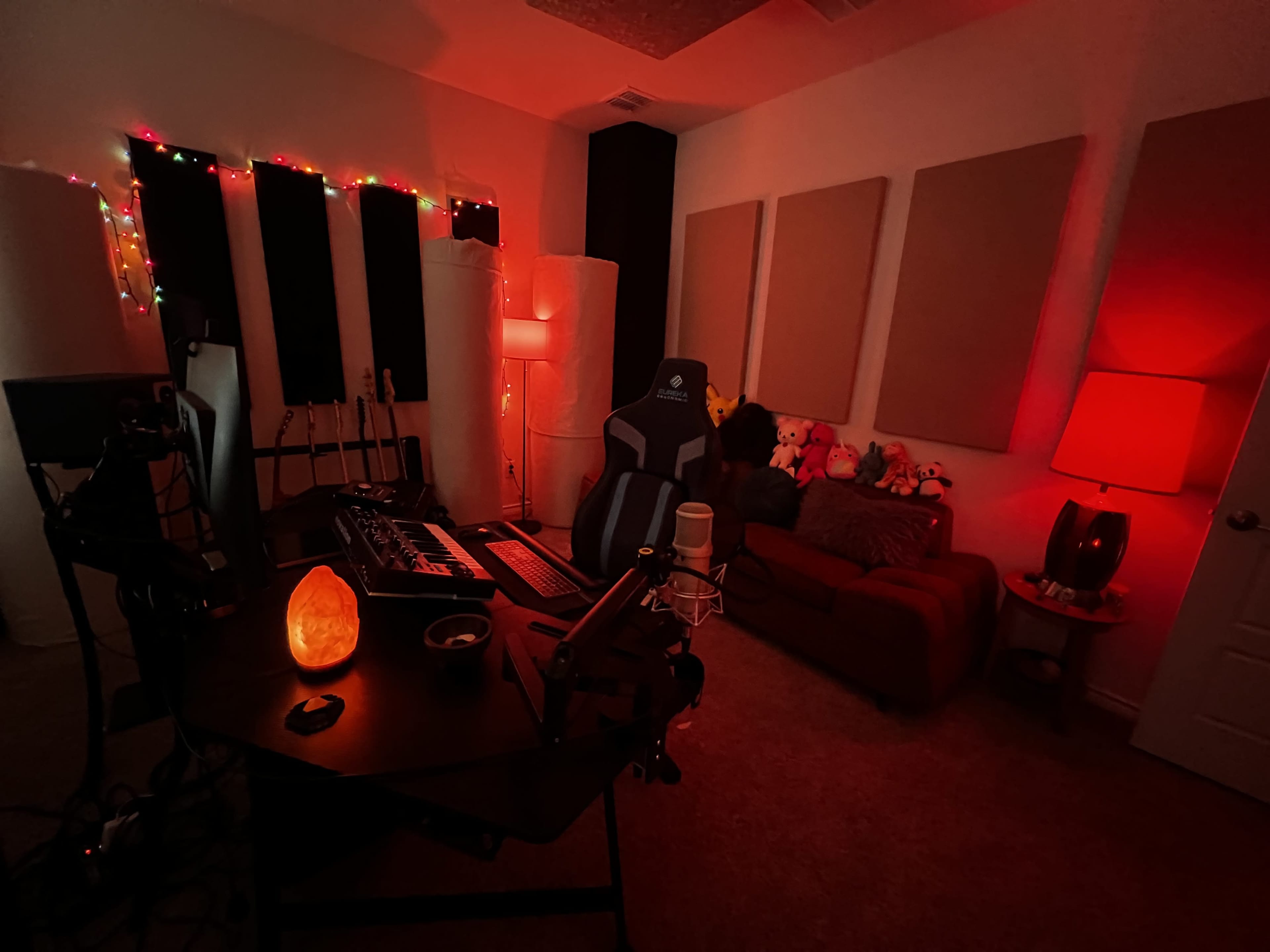 A dimly lit music studio features a desk with electronic equipment, a gaming chair, a red couch, and decorative lights lining the walls.
