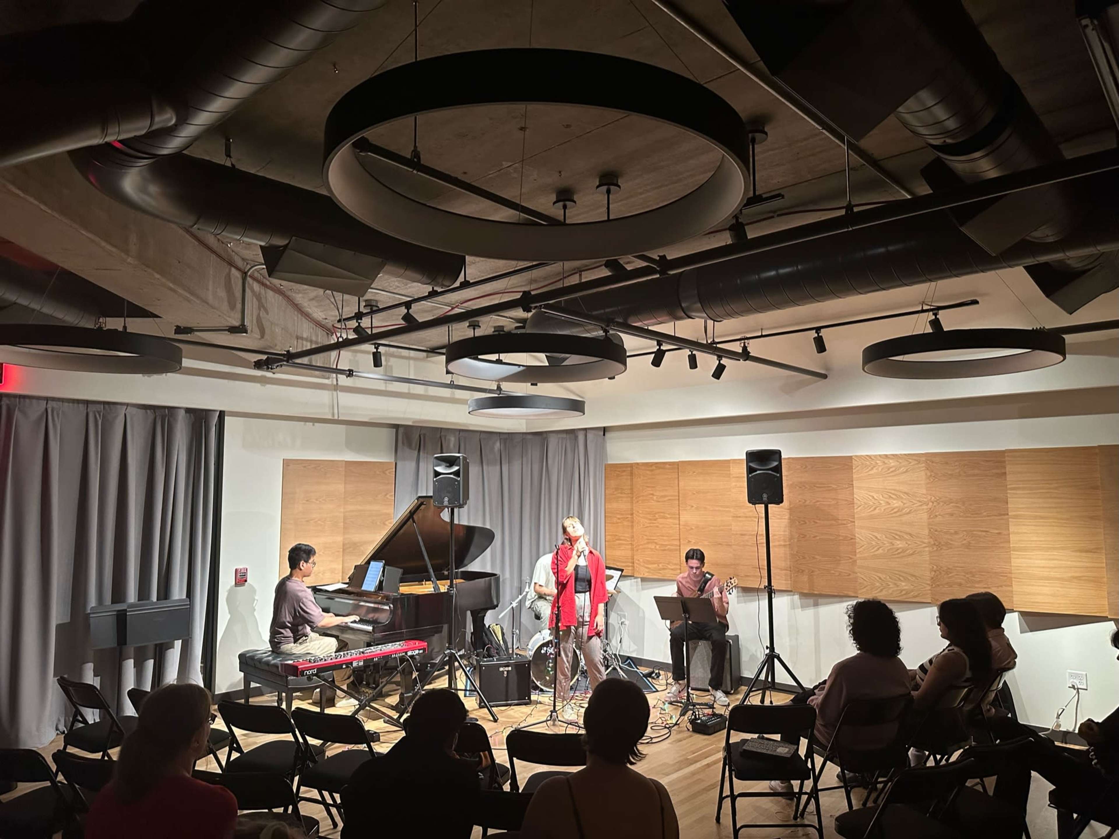A small music venue features a trio of musicians performing on stage, with a pianist, a drummer, and a vocalist in front of an audience seated in folding chairs.