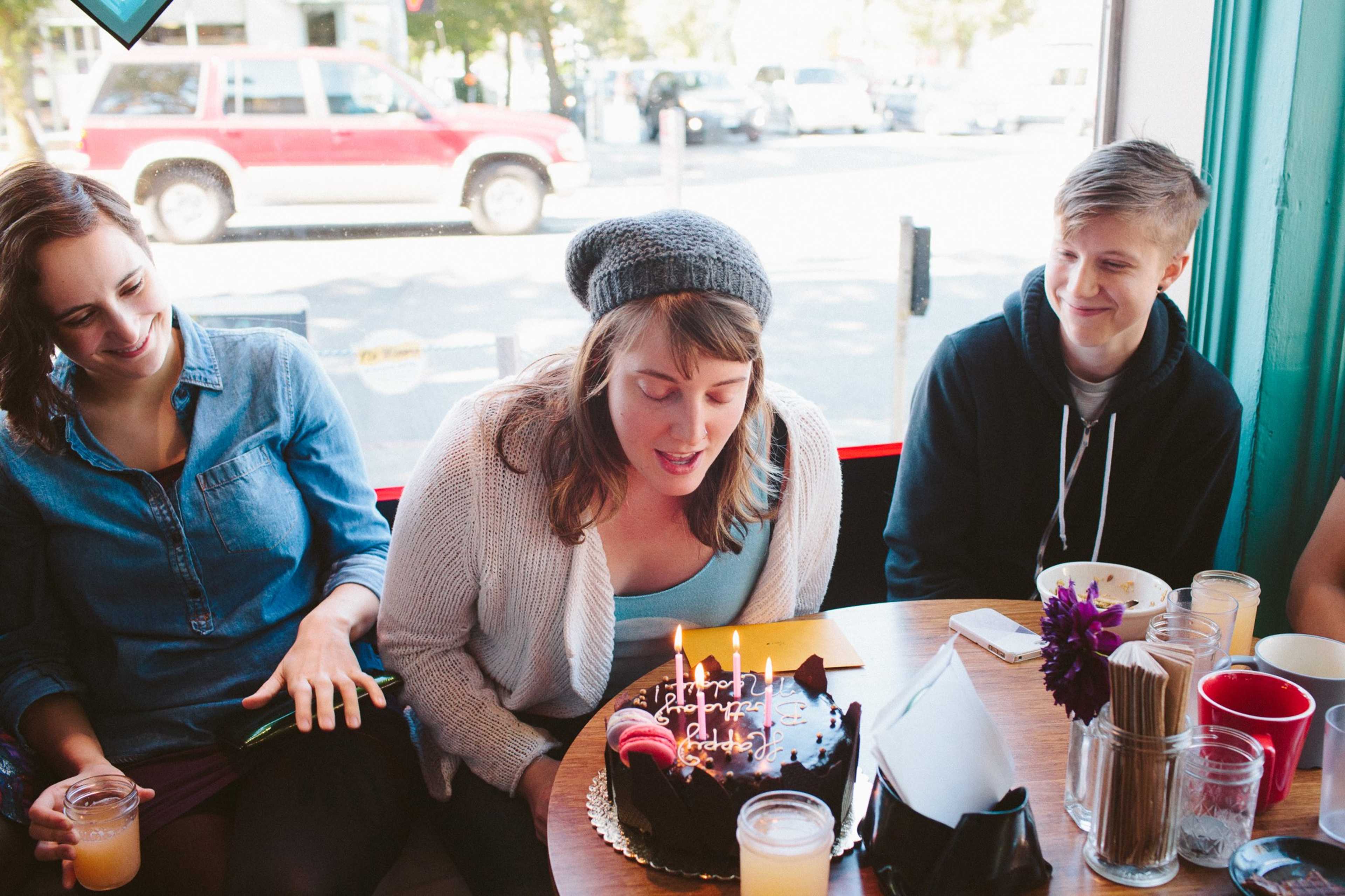 A group of friends gathers around a table with a birthday cake as one person prepares to blow out the candles.