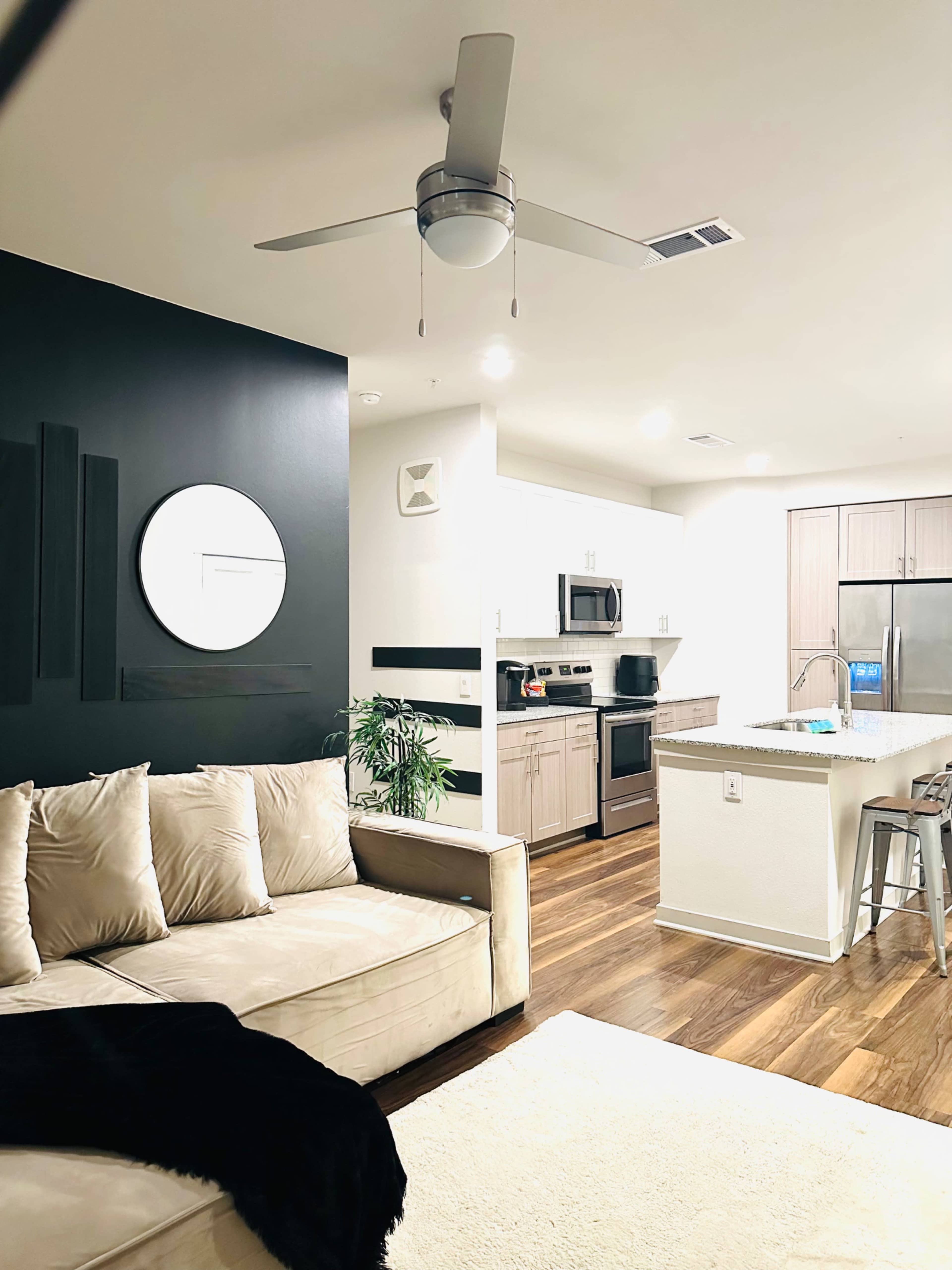 The image shows a modern living room and kitchen area featuring a beige sectional sofa, a wall-mounted round mirror, and stainless steel appliances.