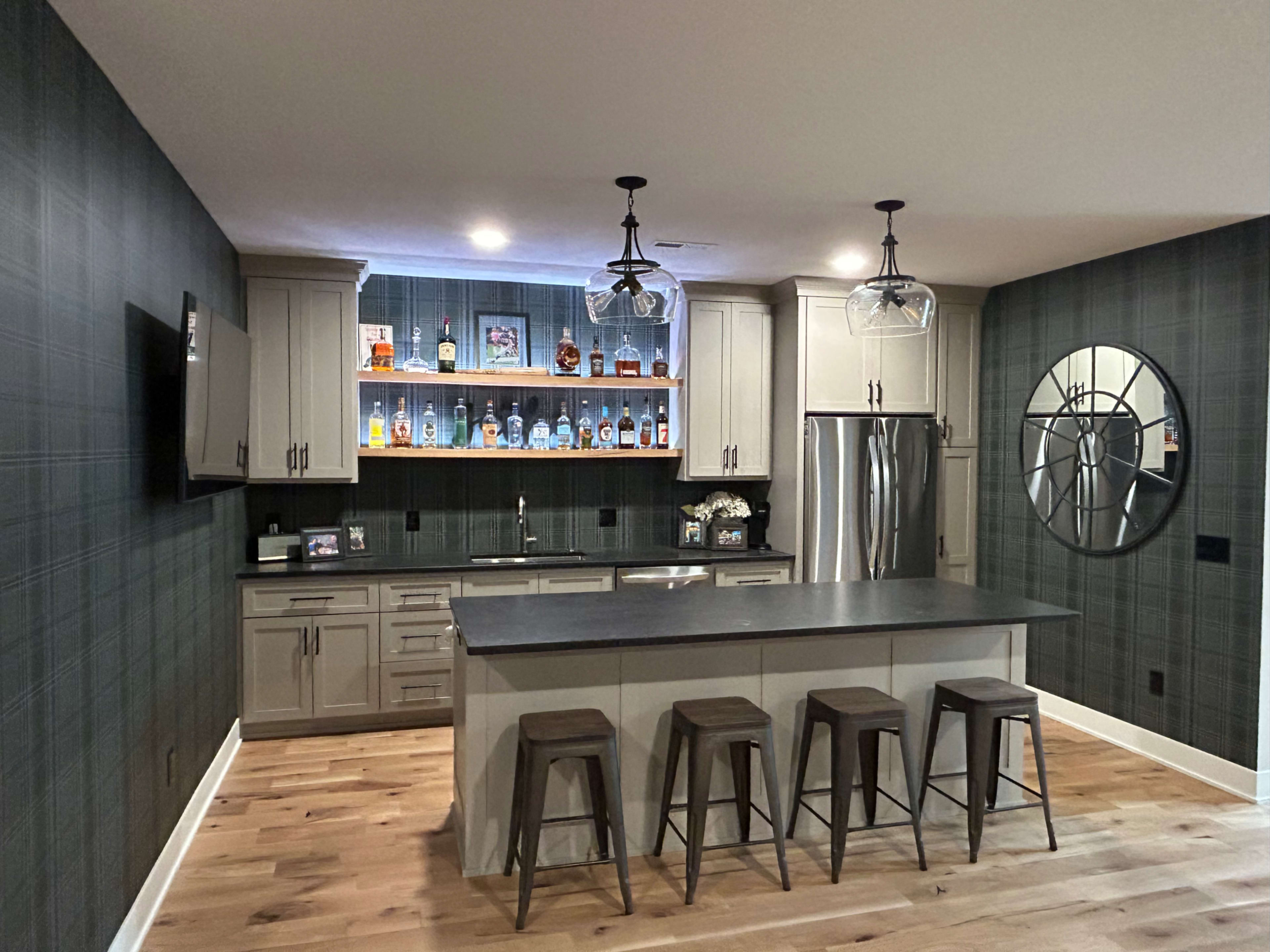 The image shows a modern kitchen with a gray color scheme, featuring a large island with stools, a wall-mounted television, and a backlit shelf displaying several bottles.