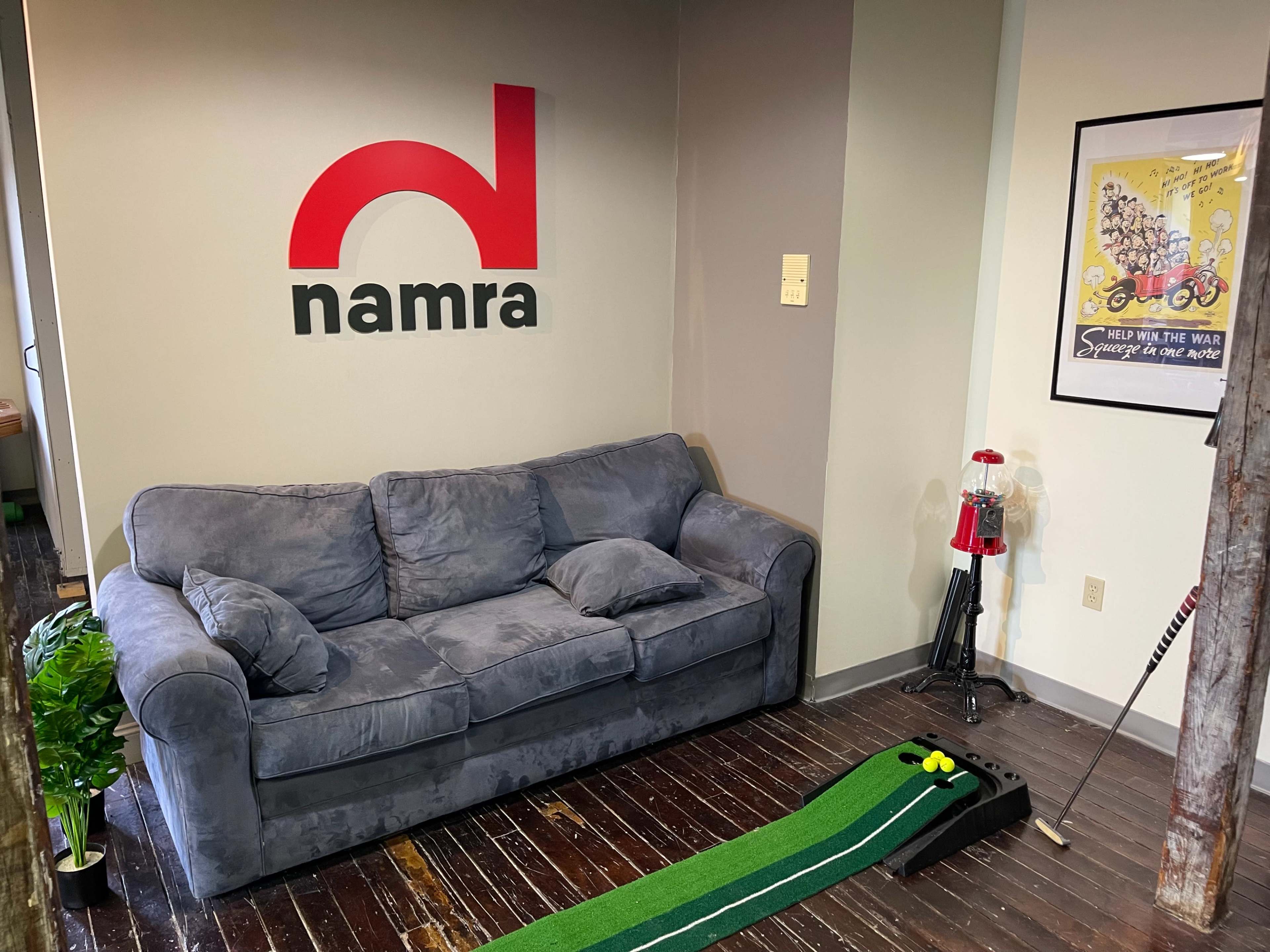 A small lounge area featuring a gray couch, a miniature putting green, and a wall with a red and black logo that reads "namra."