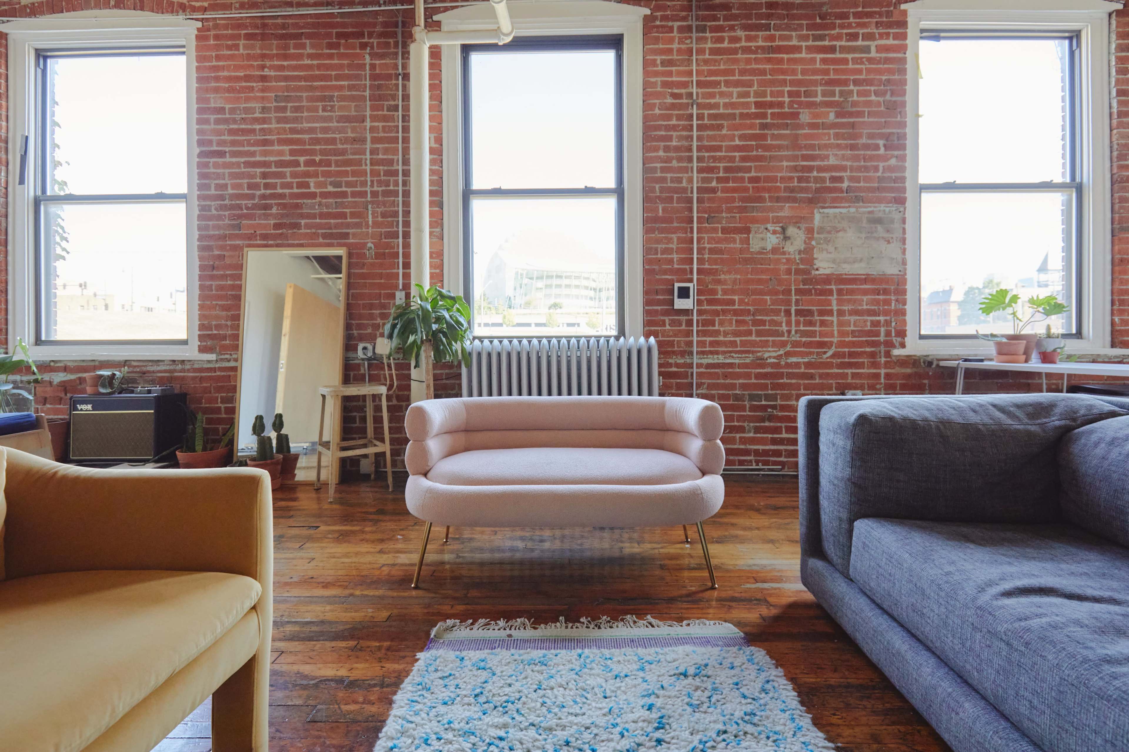 A light pink sofa is positioned in a spacious room with exposed brick walls and large windows, complemented by a few plants and a gray couch.