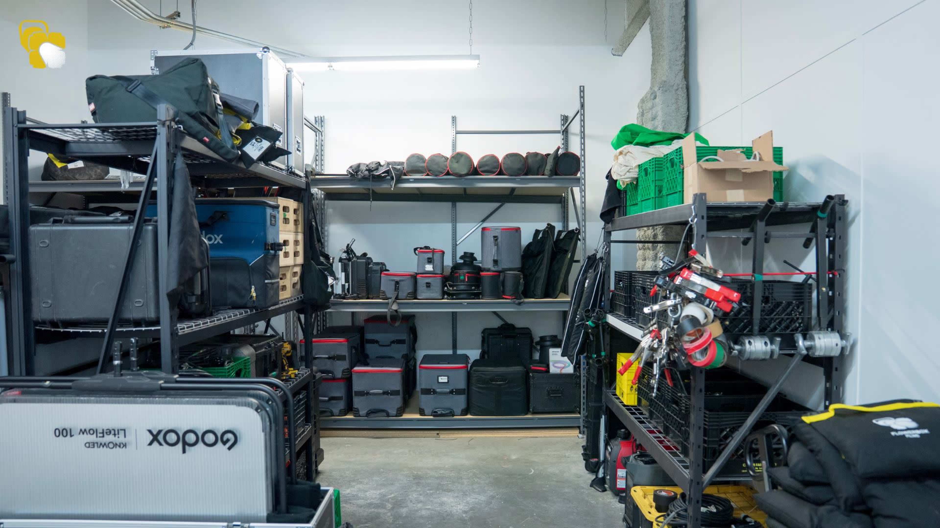 The image shows a storage area with metal shelves filled with various equipment cases, bags, and tools organized neatly along the walls.