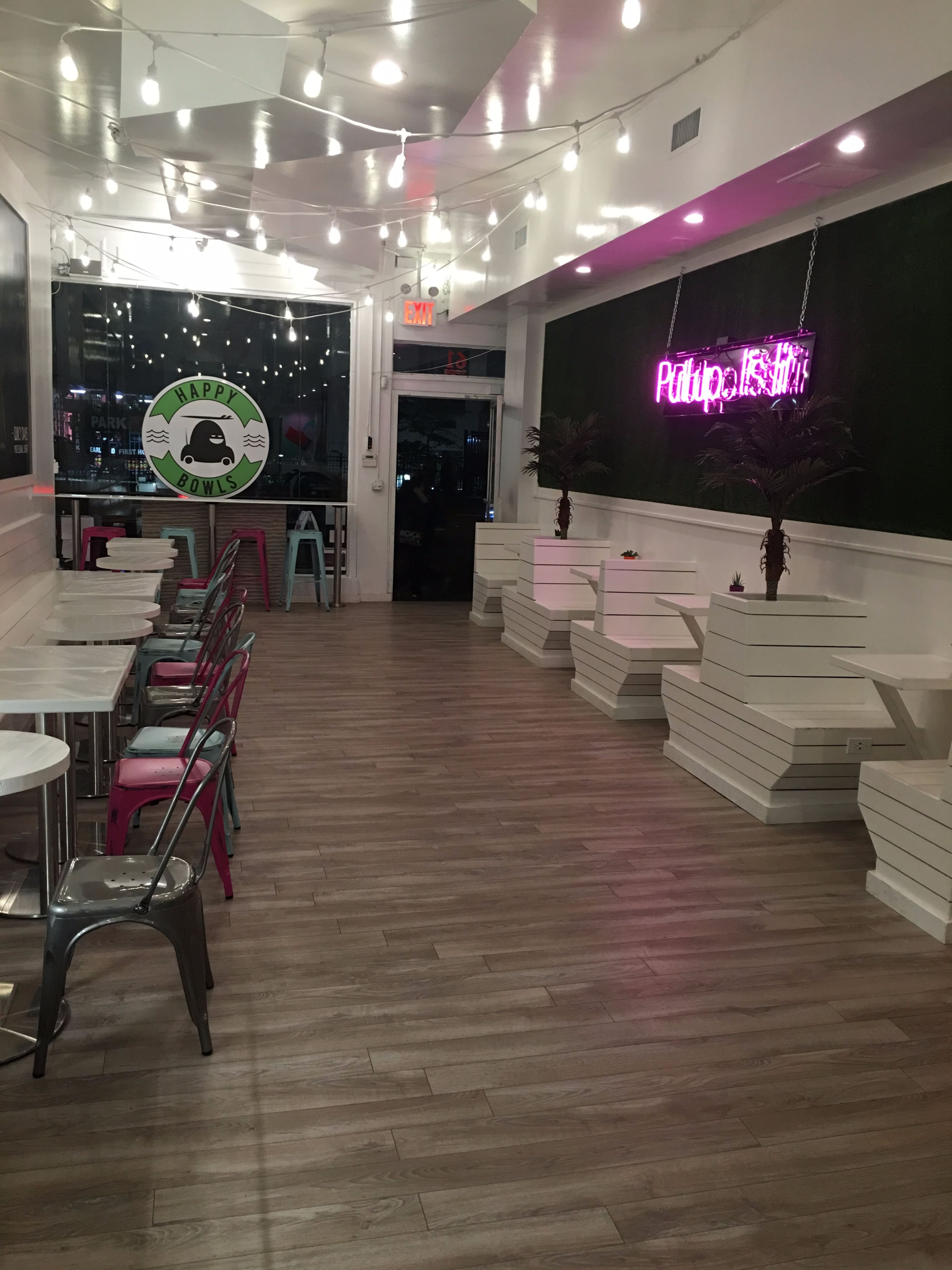 The image shows a brightly lit interior of a cafe with wooden flooring, minimalistic white furniture, and neon signs on the walls.