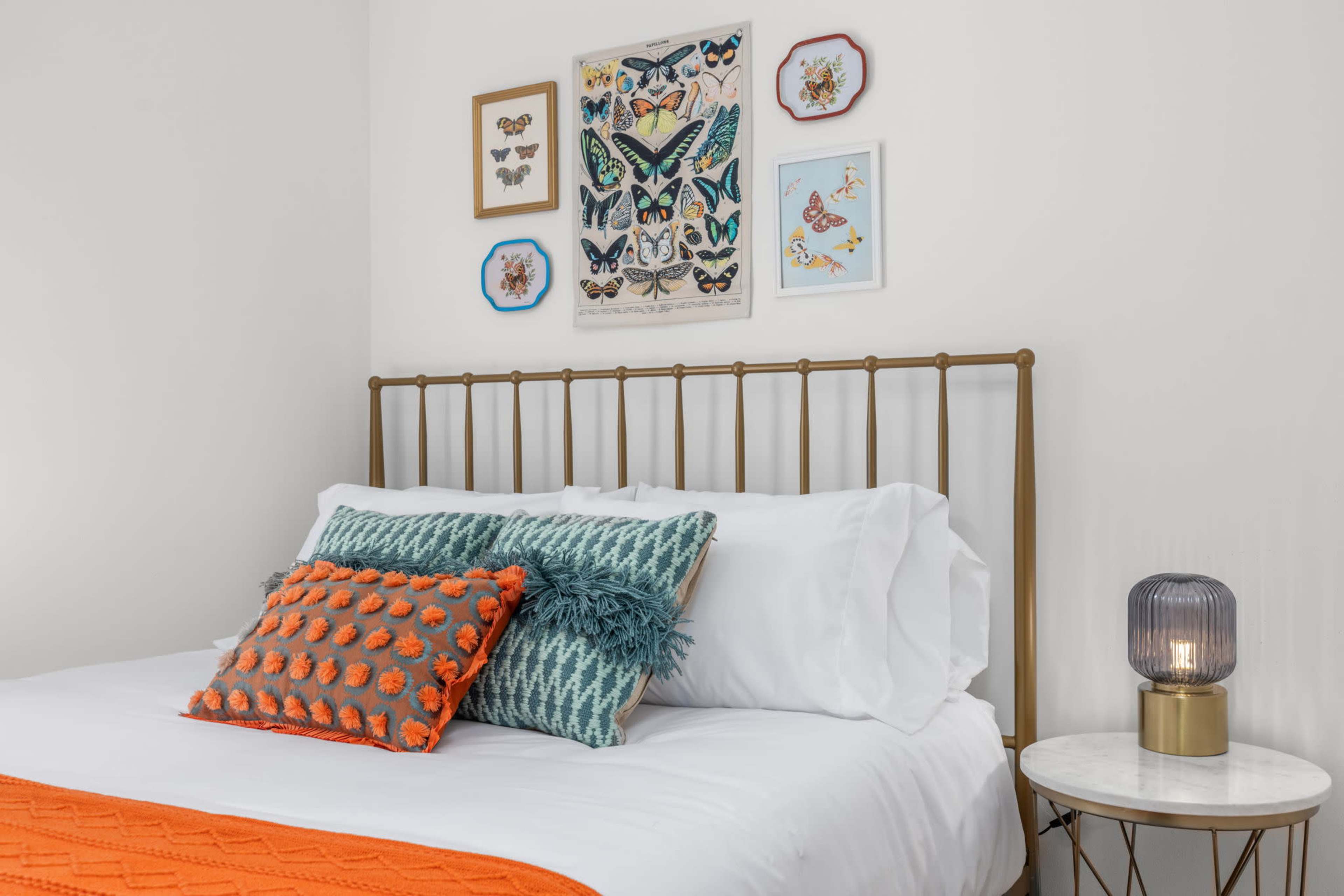 The image shows a neatly made bed with decorative pillows, a patterned orange throw blanket, and framed artwork of butterflies on the wall above.
