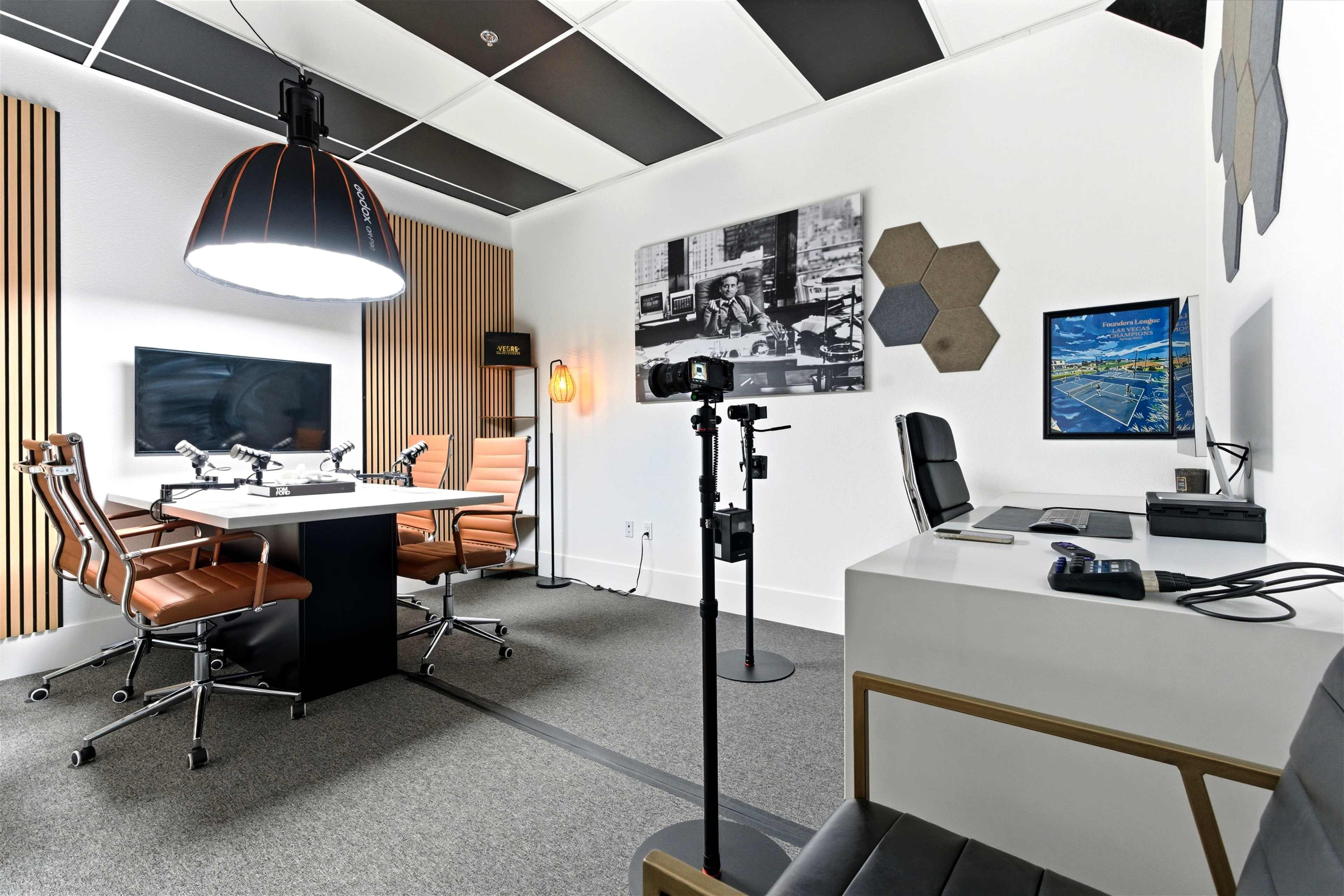 The image shows a modern conference room equipped with a rectangular table, ergonomic chairs, a wall-mounted screen, and a camera setup on a tripod.