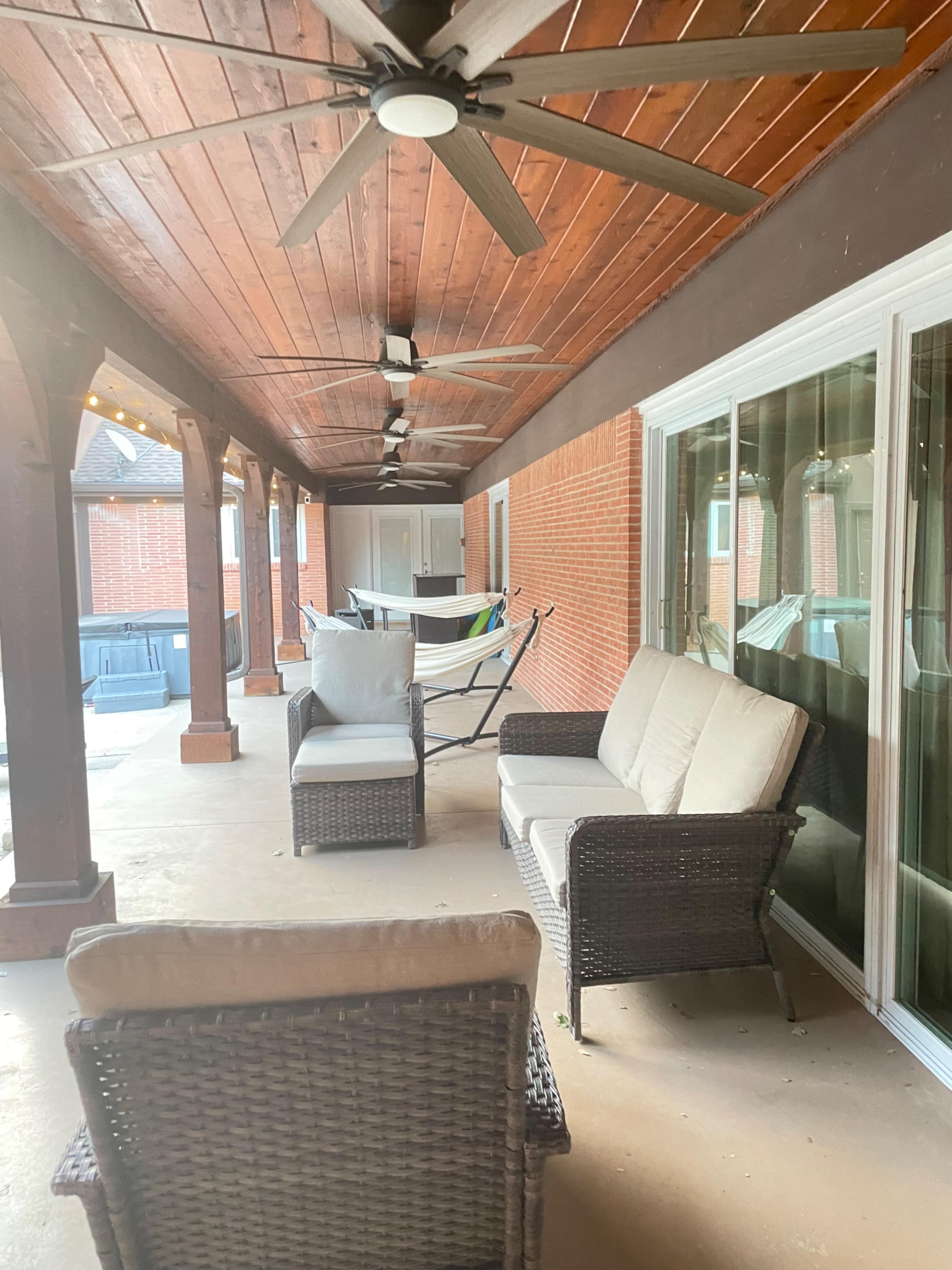 The image shows a covered patio featuring wicker furniture, a ceiling fan, and a hammock in the background.