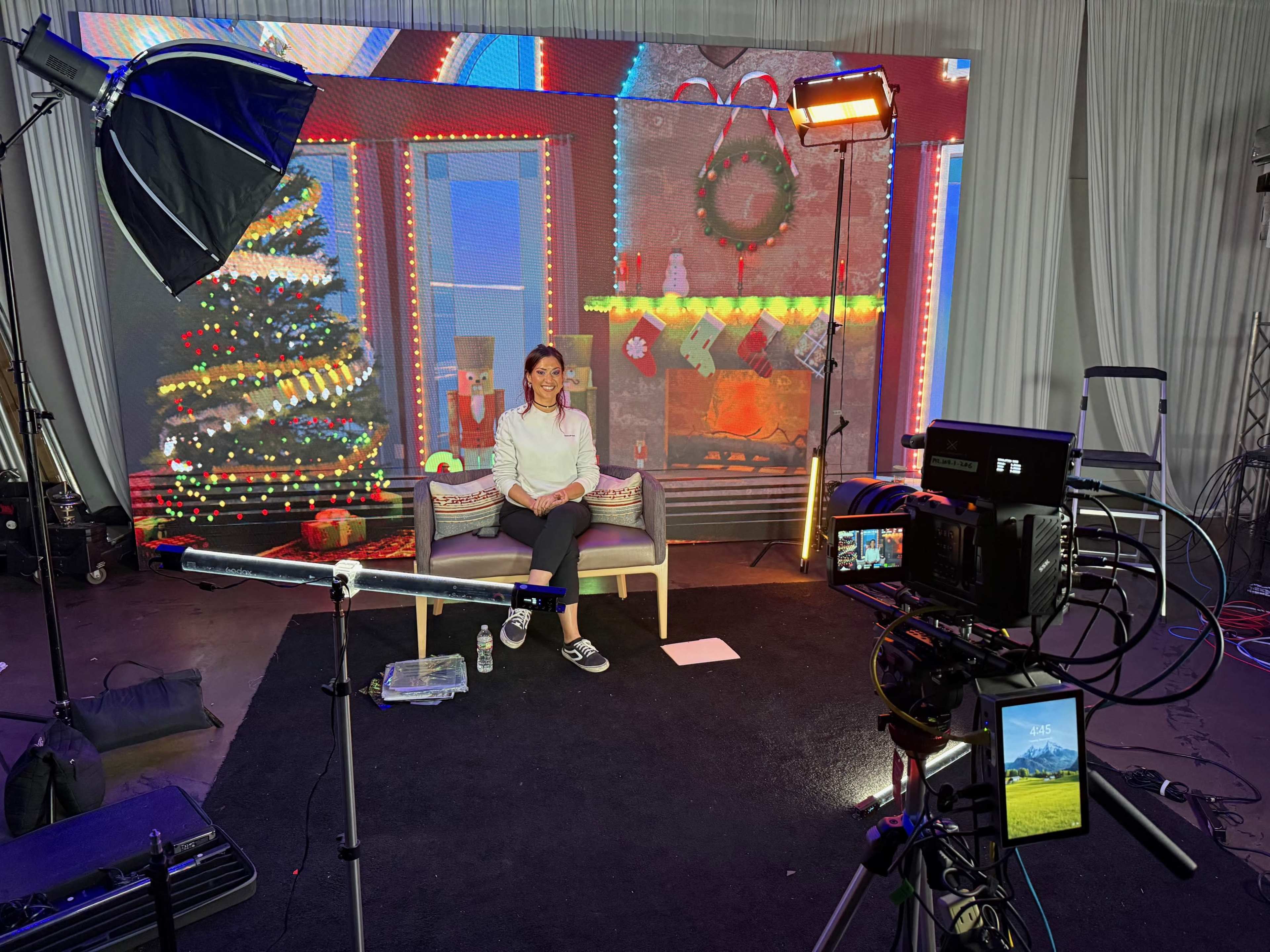 A person is seated on a couch in a studio decorated with holiday-themed lights and a backdrop featuring a Christmas tree and a fireplace.
