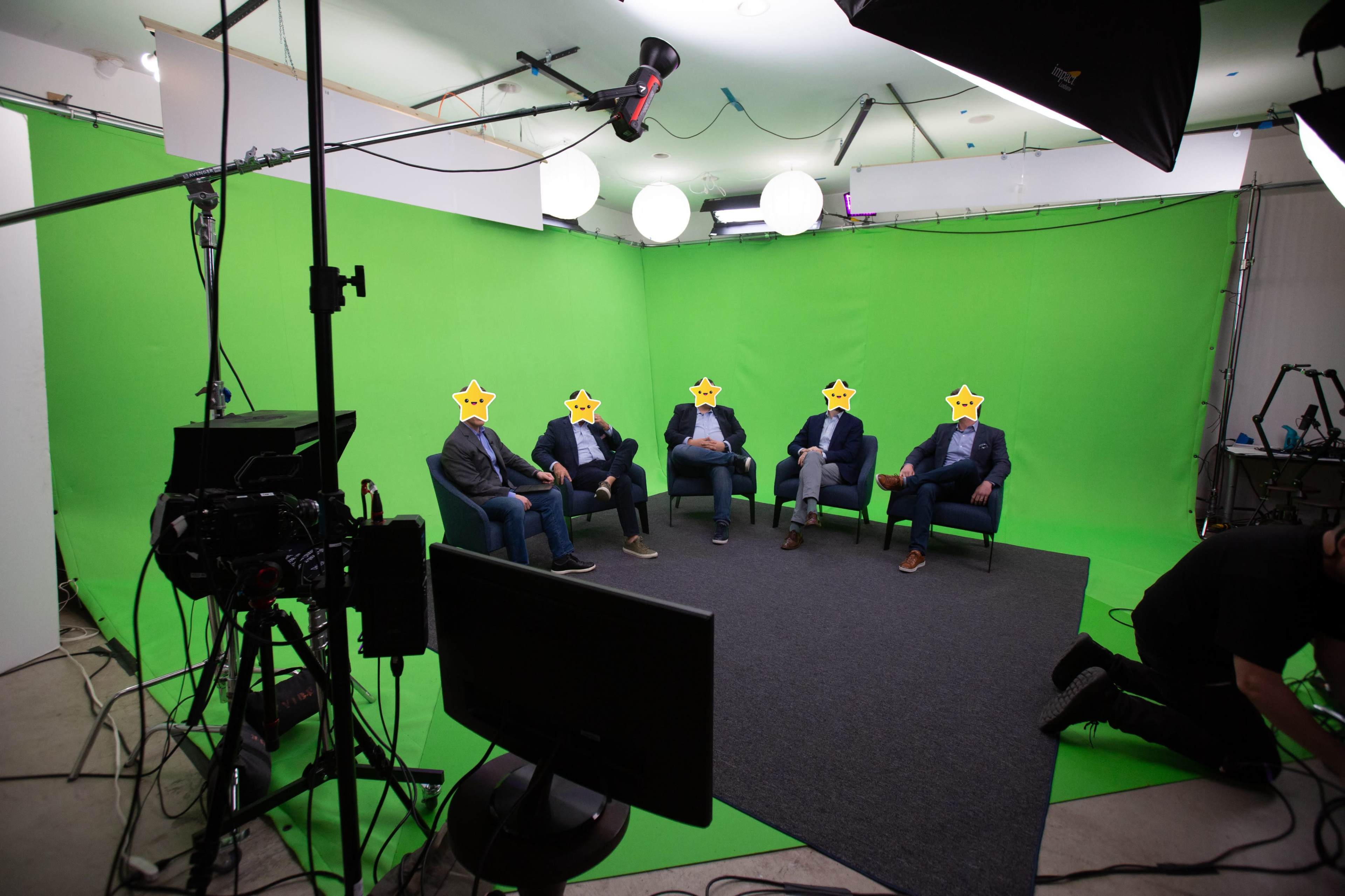 A group of five people sits on chairs in front of a green screen, surrounded by camera equipment and studio lighting.