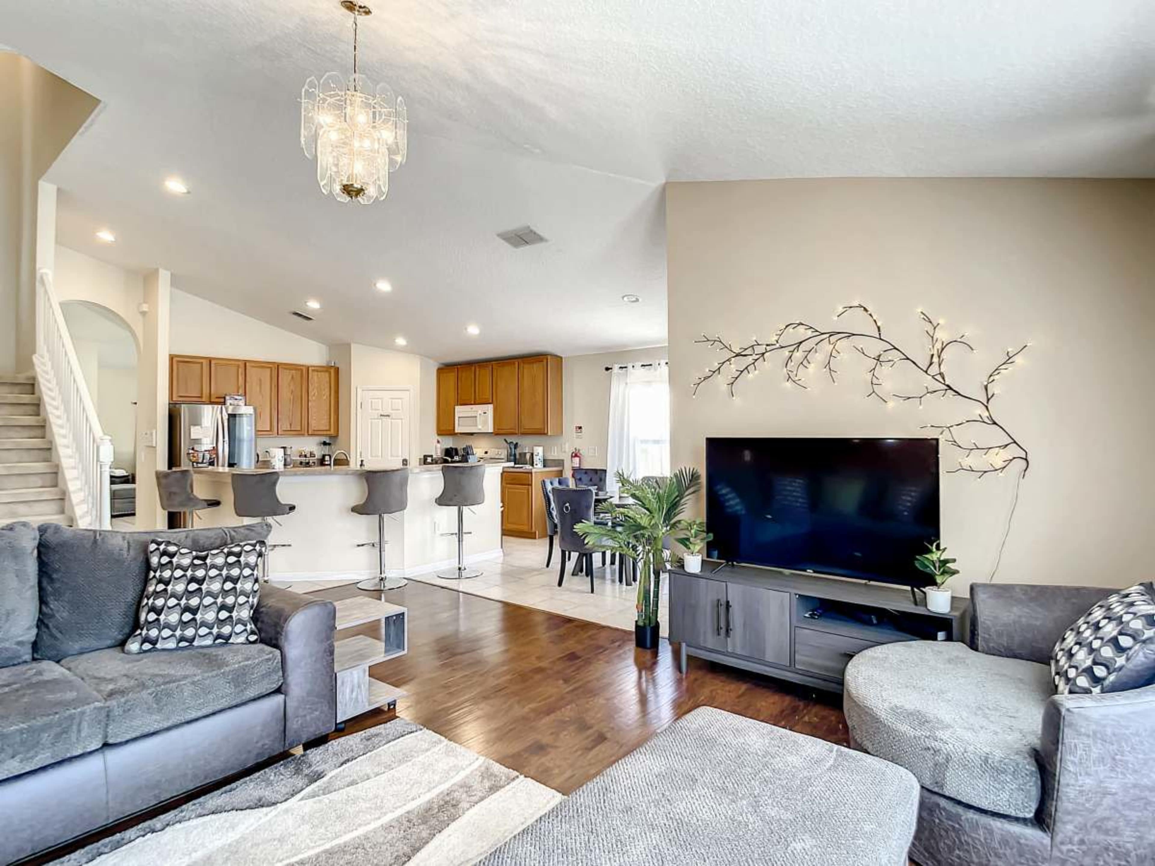 The image shows a modern living room with a large television, comfy seating, and a kitchen visible in the background.