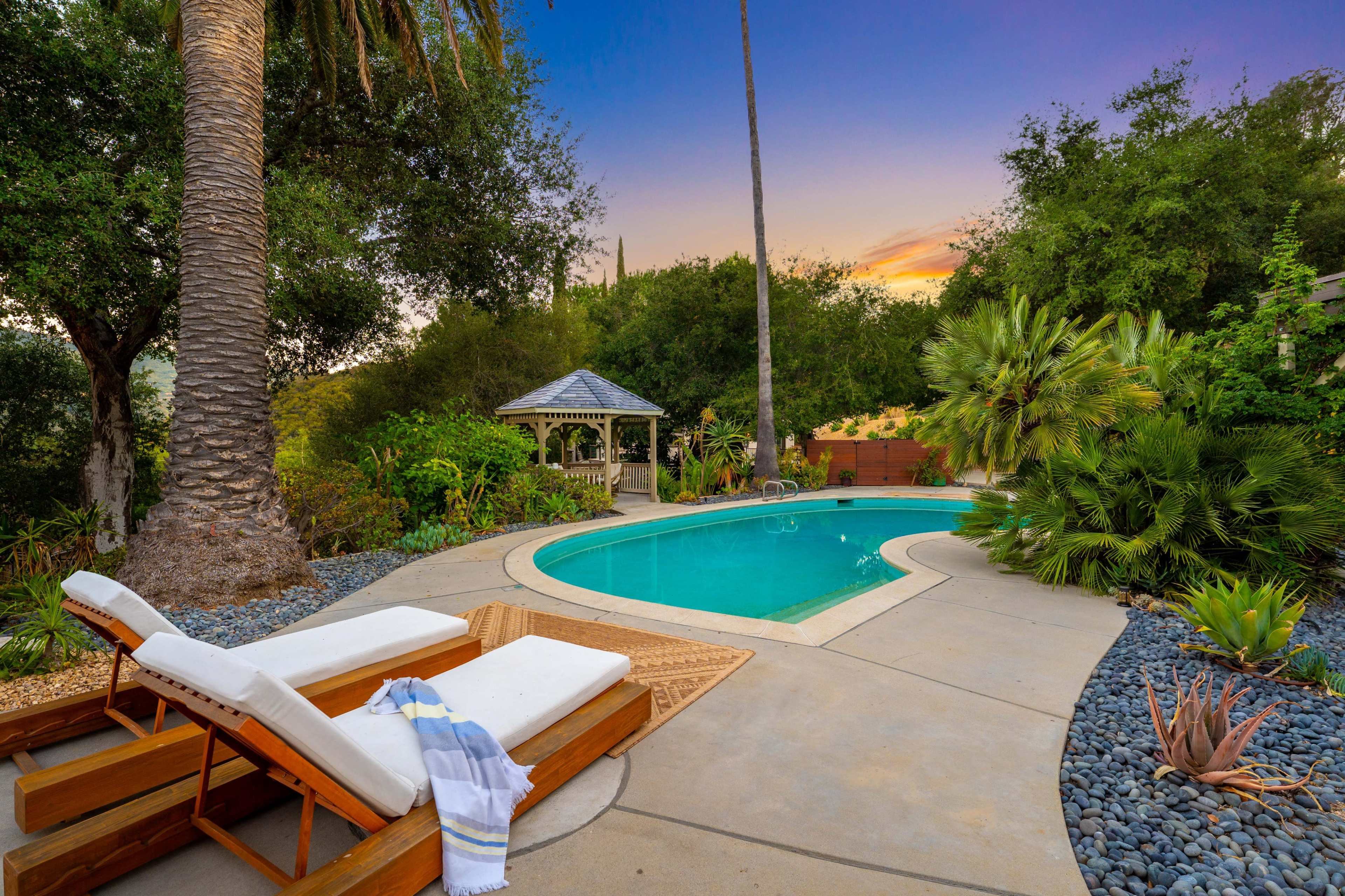 The image shows a backyard with a swimming pool surrounded by tropical plants and a gazebo, with lounge chairs placed nearby.