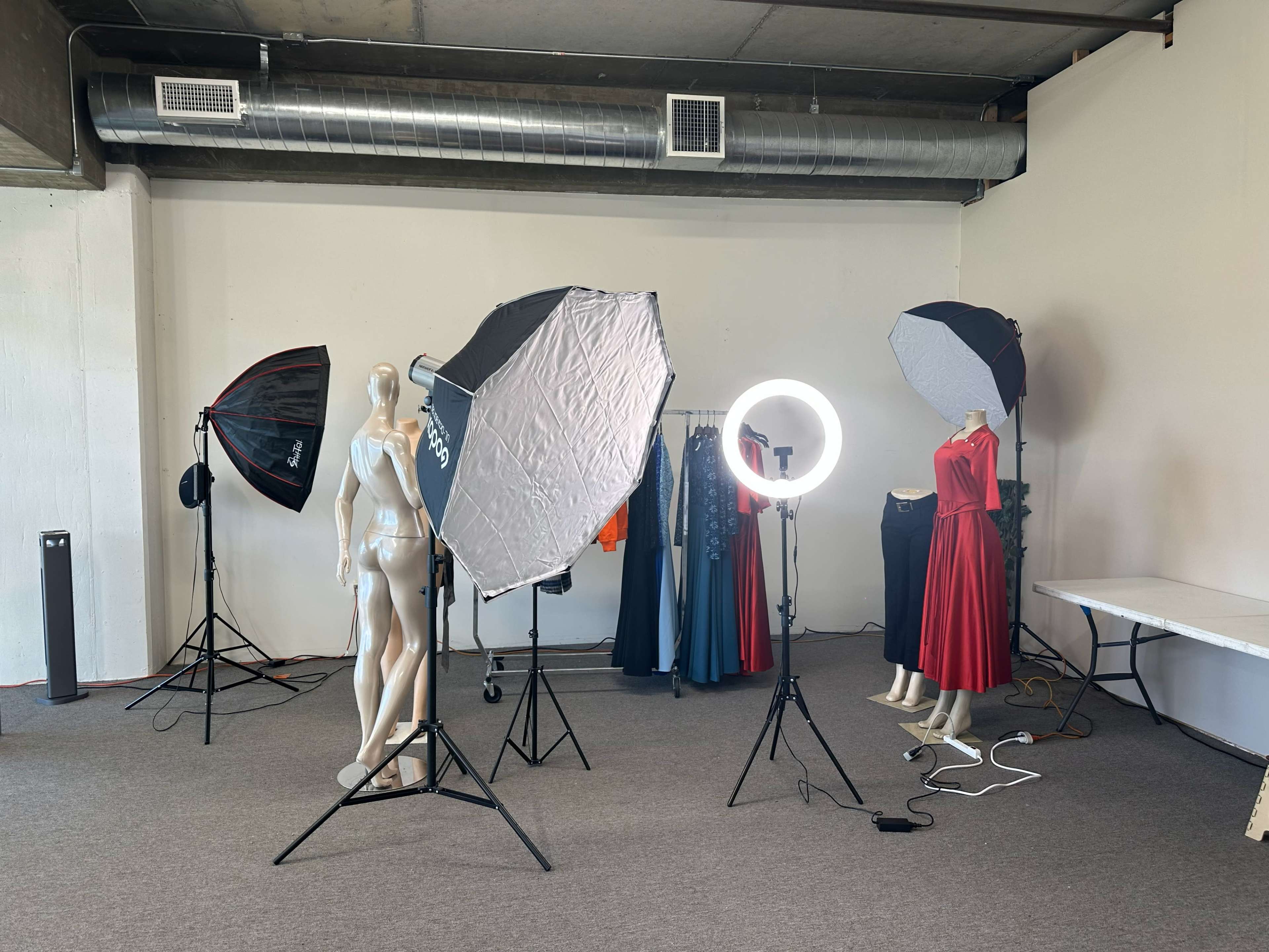 The image shows a photography studio setup featuring a mannequin, two garment racks with dresses, and various lighting equipment, including softboxes and a ring light.
