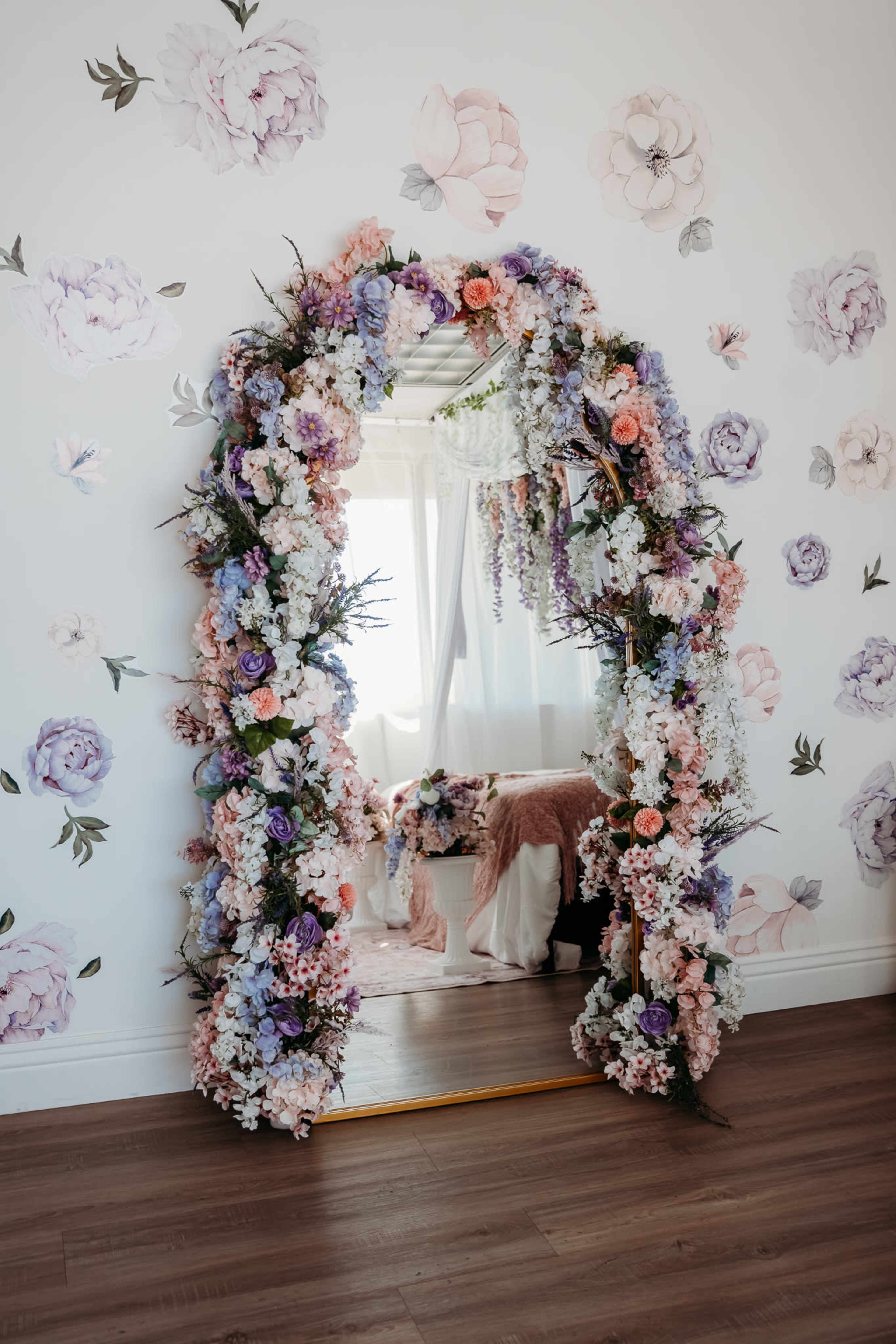 A large mirror is framed with a floral arrangement in shades of purple, pink, and white, set against a wall adorned with pastel floral wallpaper.