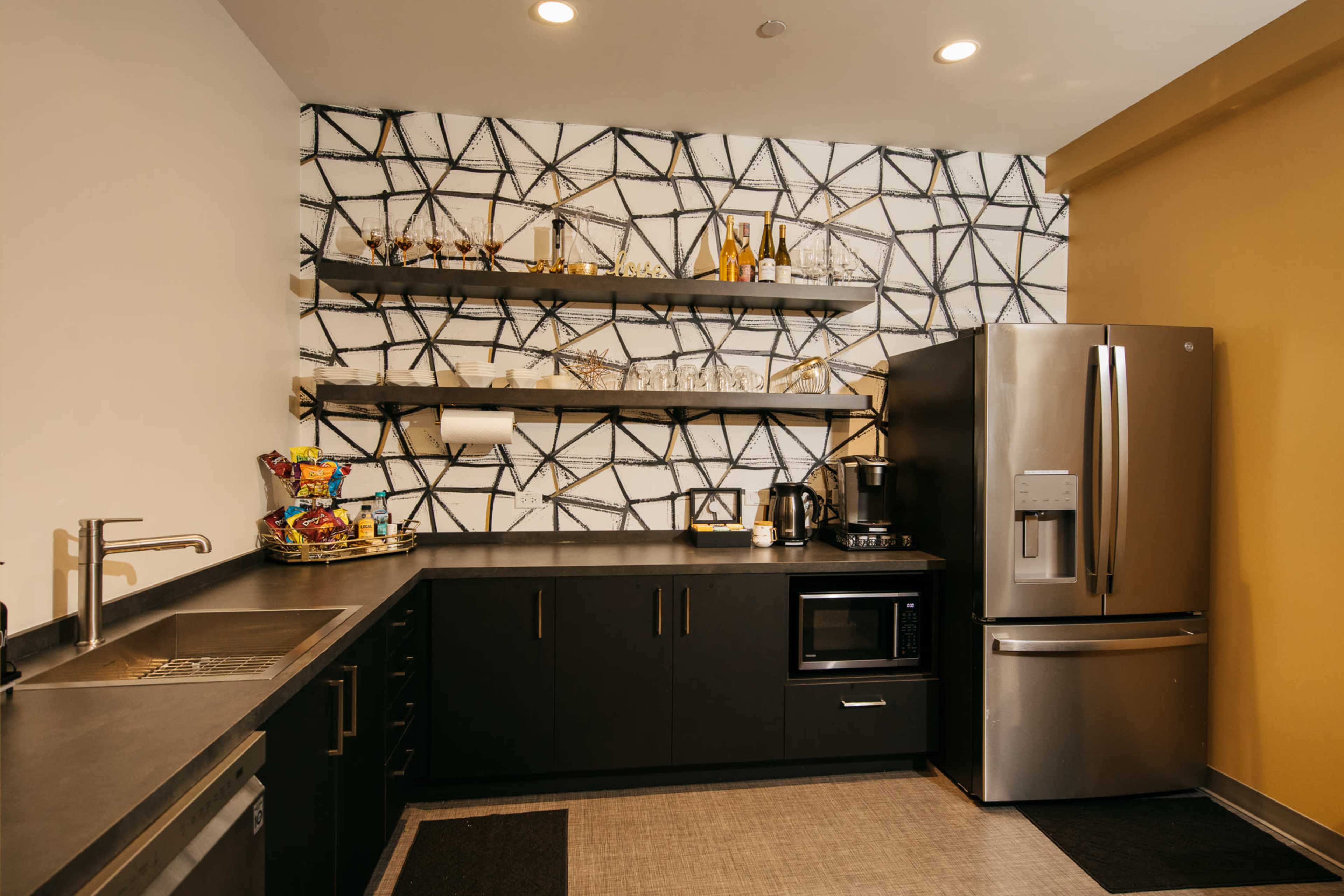A modern kitchen with black cabinets, a stainless steel refrigerator, and open shelves displaying glassware and kitchen items against a patterned wall.