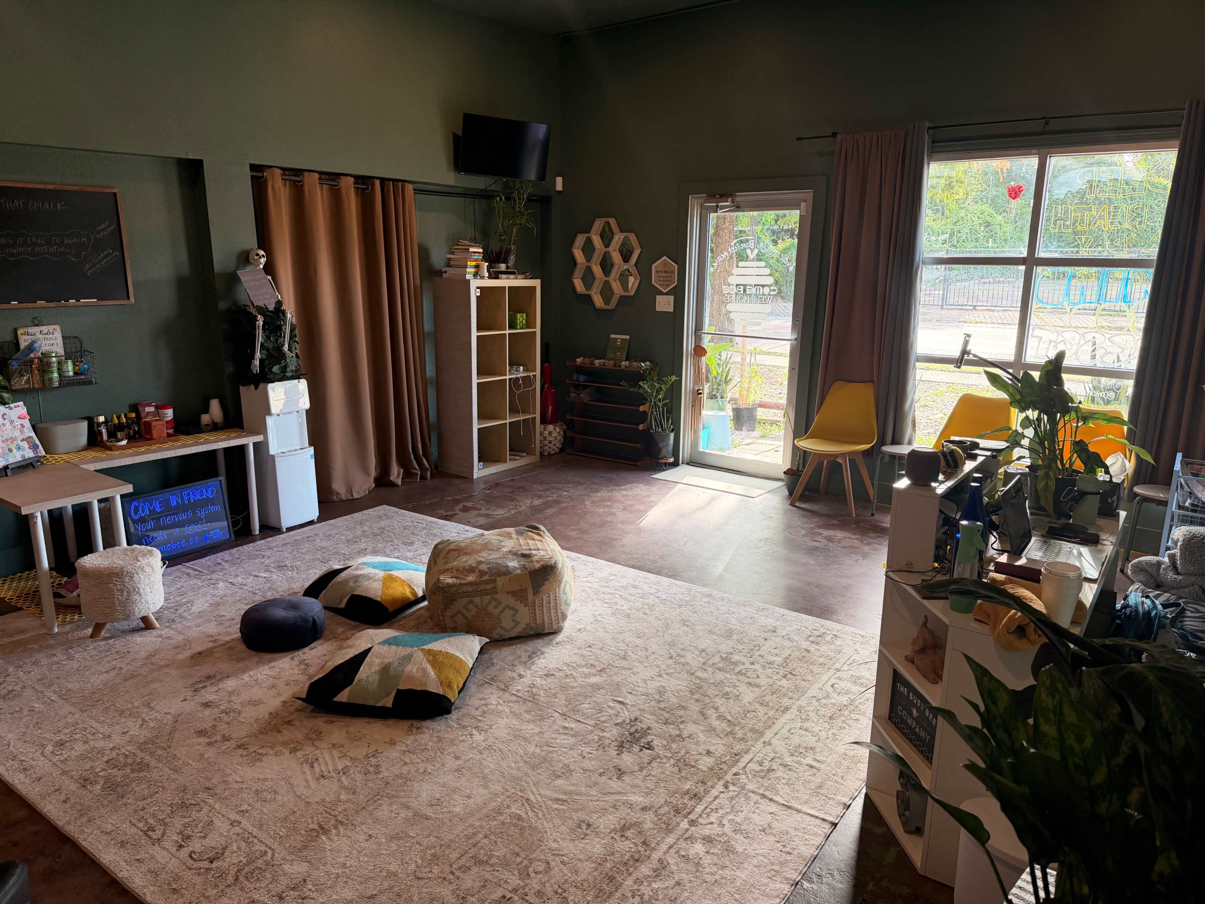 The image shows a spacious, well-lit room with a large rug, a seating area with cushions, shelves, and a doorway leading to an outdoor space.
