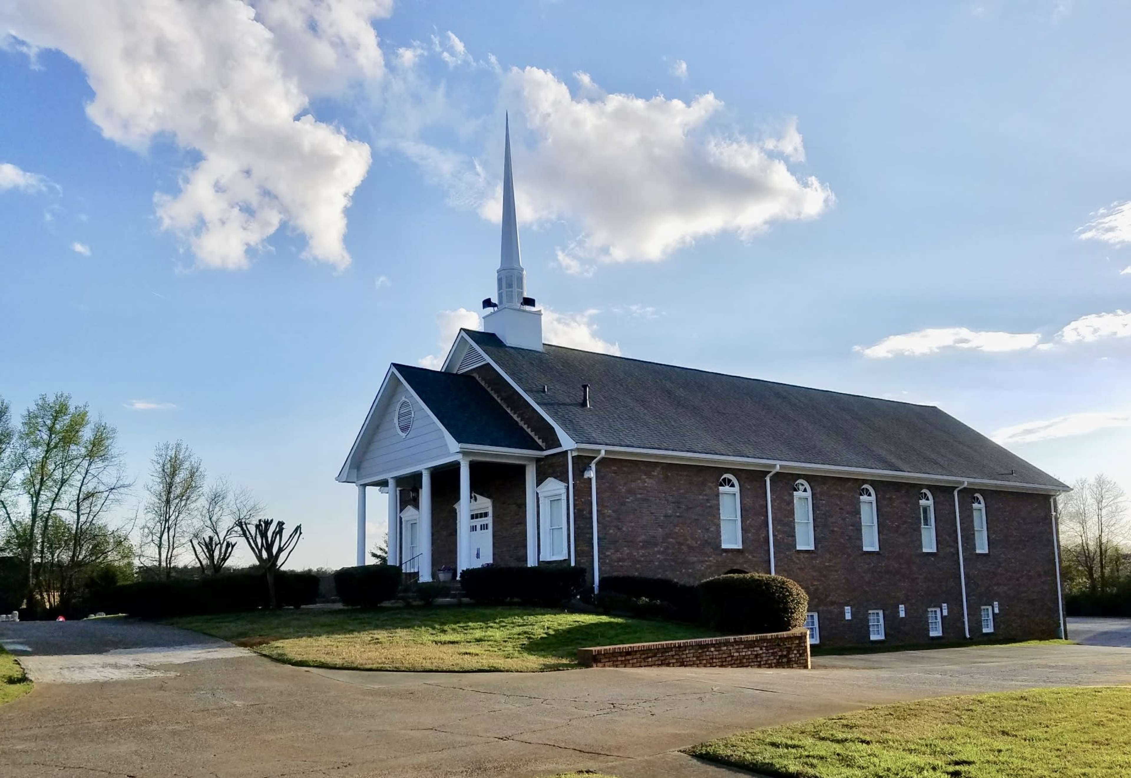 A brick church building with a steeple stands at the end of a paved driveway under a blue sky with clouds.