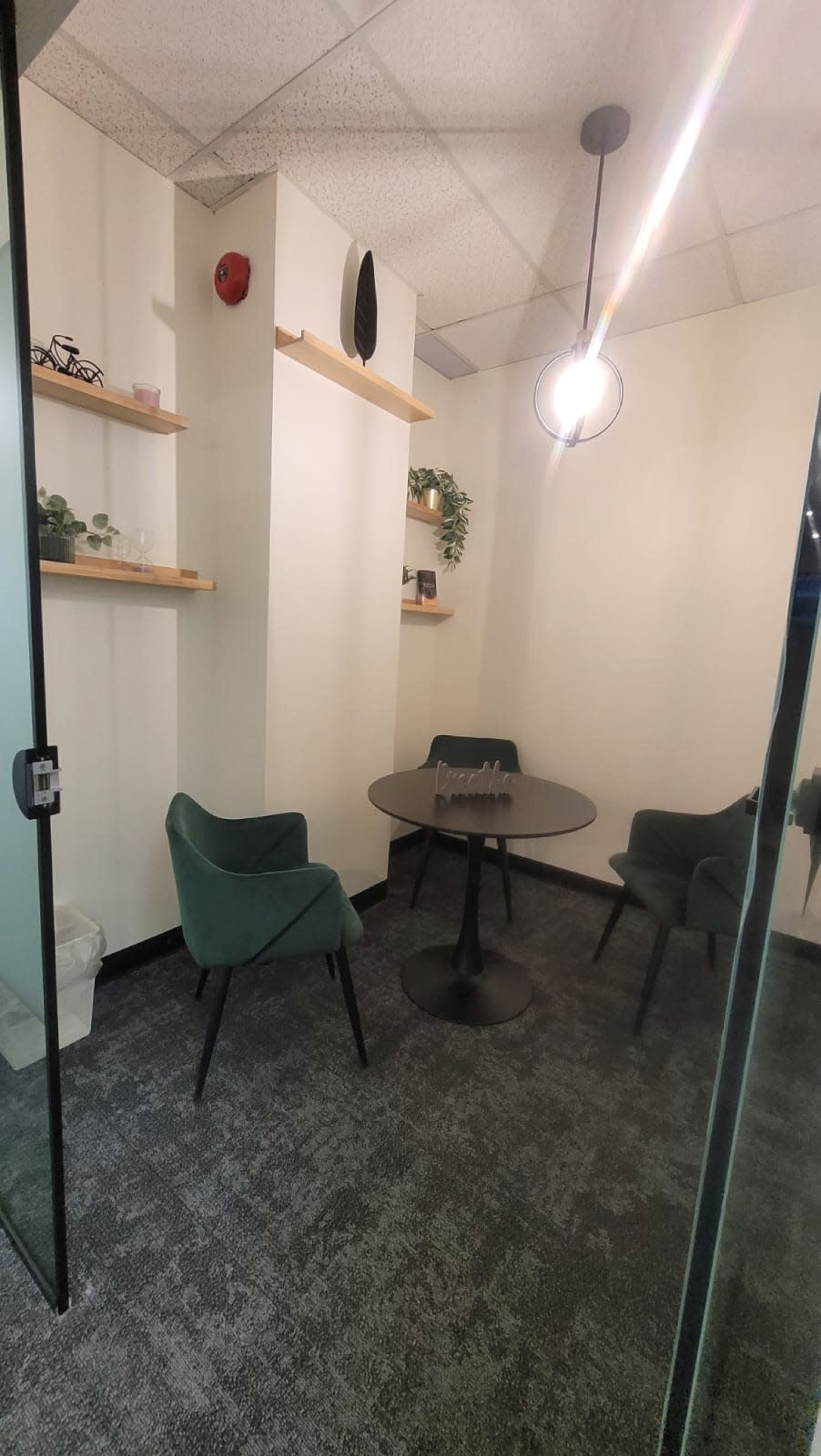 A small meeting room features a round black table surrounded by three green chairs, with shelves displaying plants and decorative items on the wall.