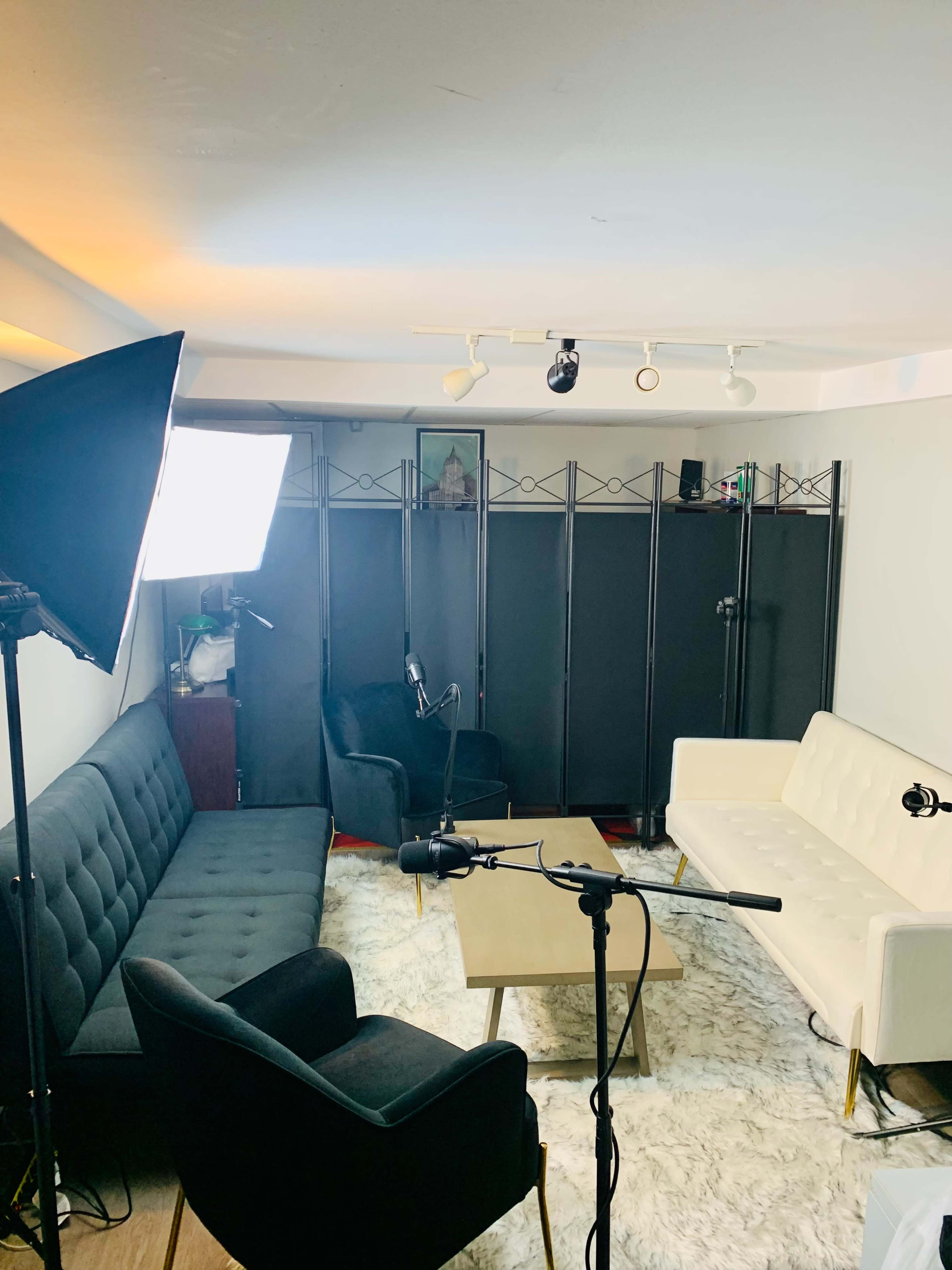 A small, well-lit recording studio with two sofas, a coffee table, and microphones set up in front of a black folding screen.