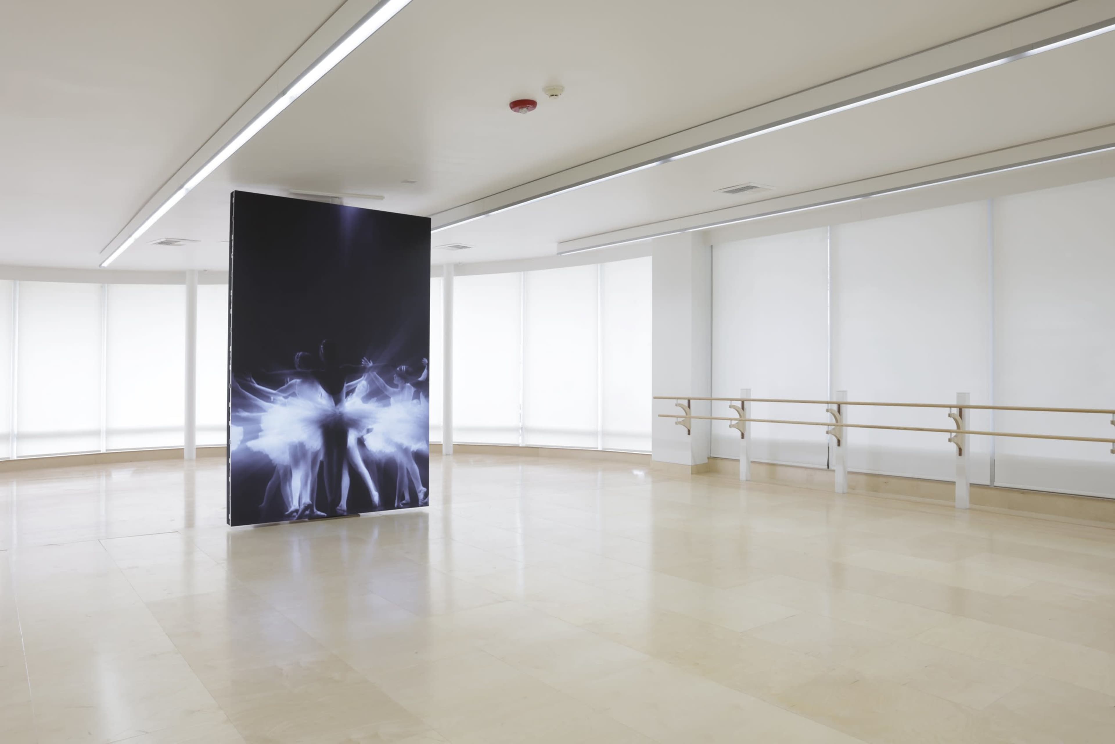 A spacious, minimalist dance studio with large windows and a central display panel featuring a graphic design.