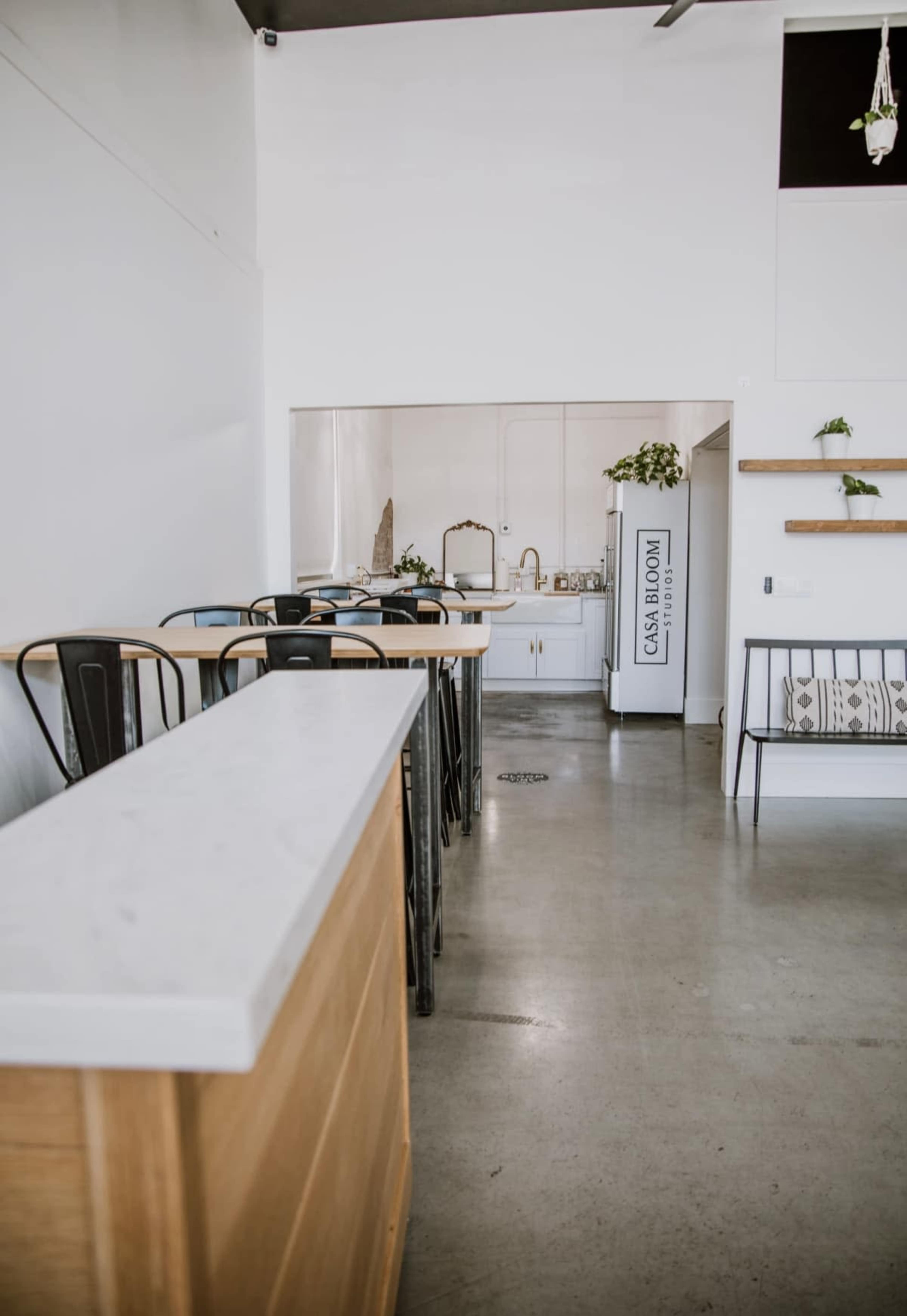 The image shows a modern, minimalistic café interior with a long bar counter, metal chairs, and a kitchen area visible in the background.