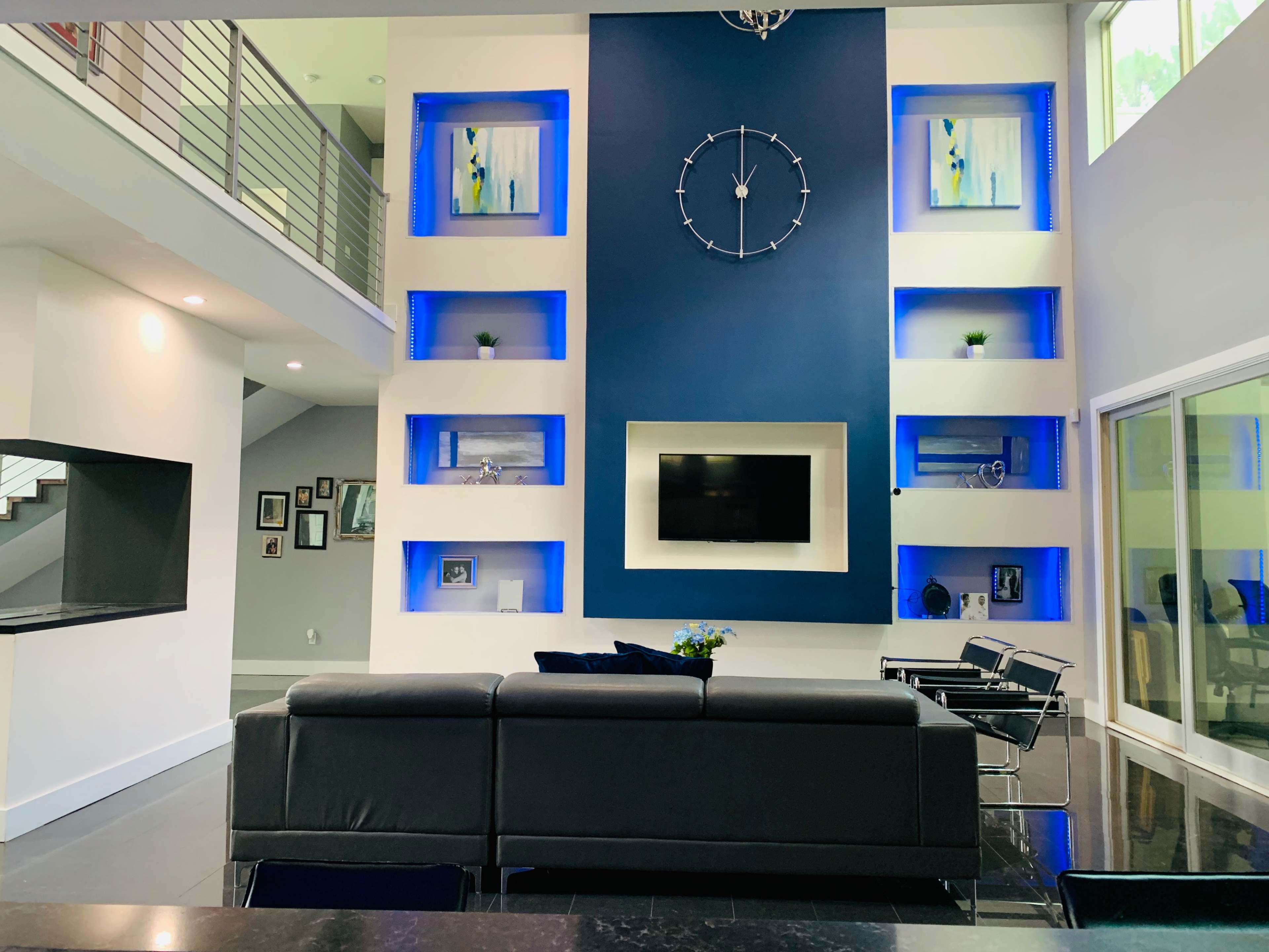 The interior of a modern living room features a dark gray sectional sofa facing a wall with blue backlighting, a clock centerpiece, and a mounted television.