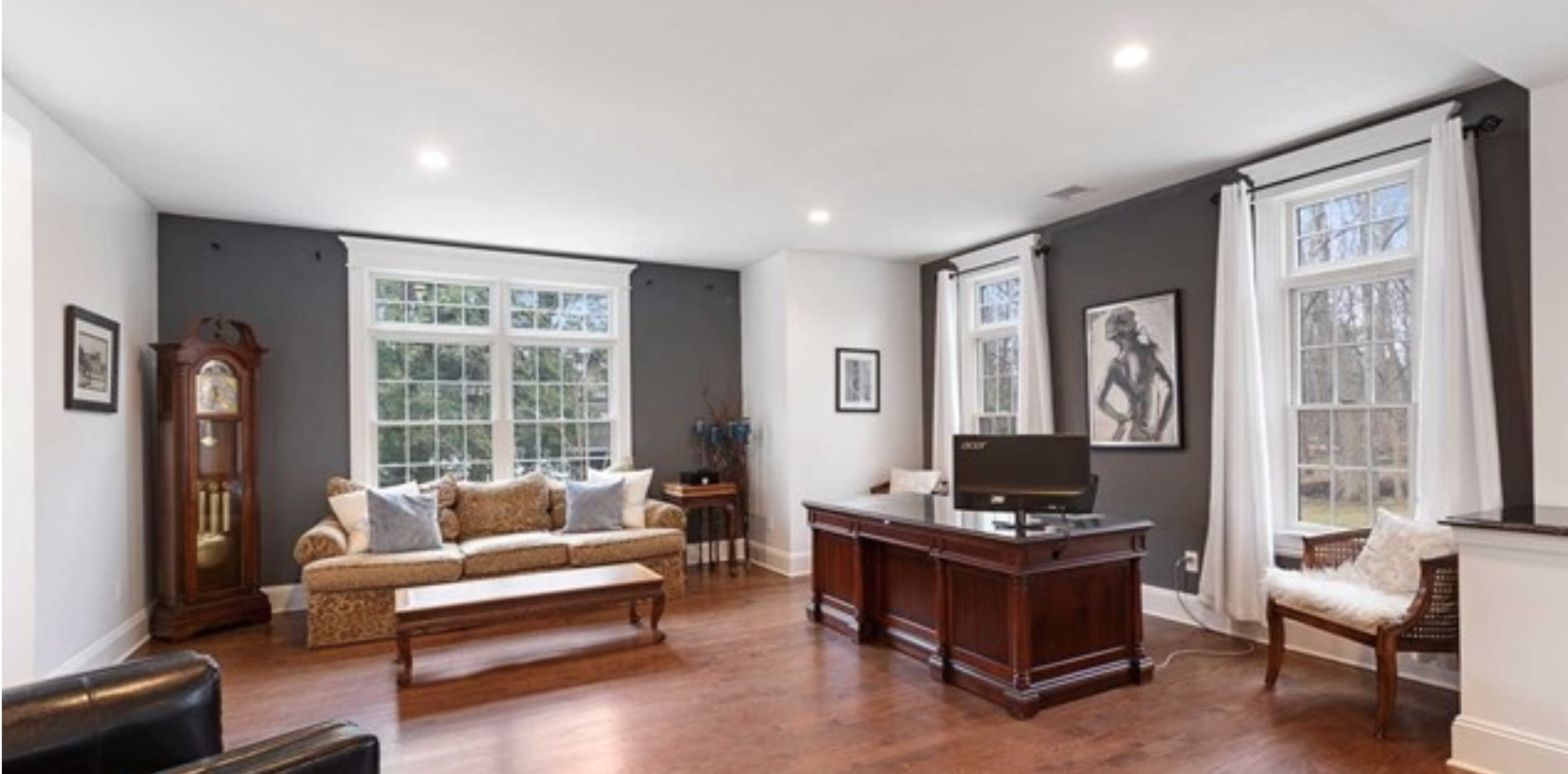 A spacious home office featuring a wooden desk, a comfortable couch, and large windows with white curtains.
