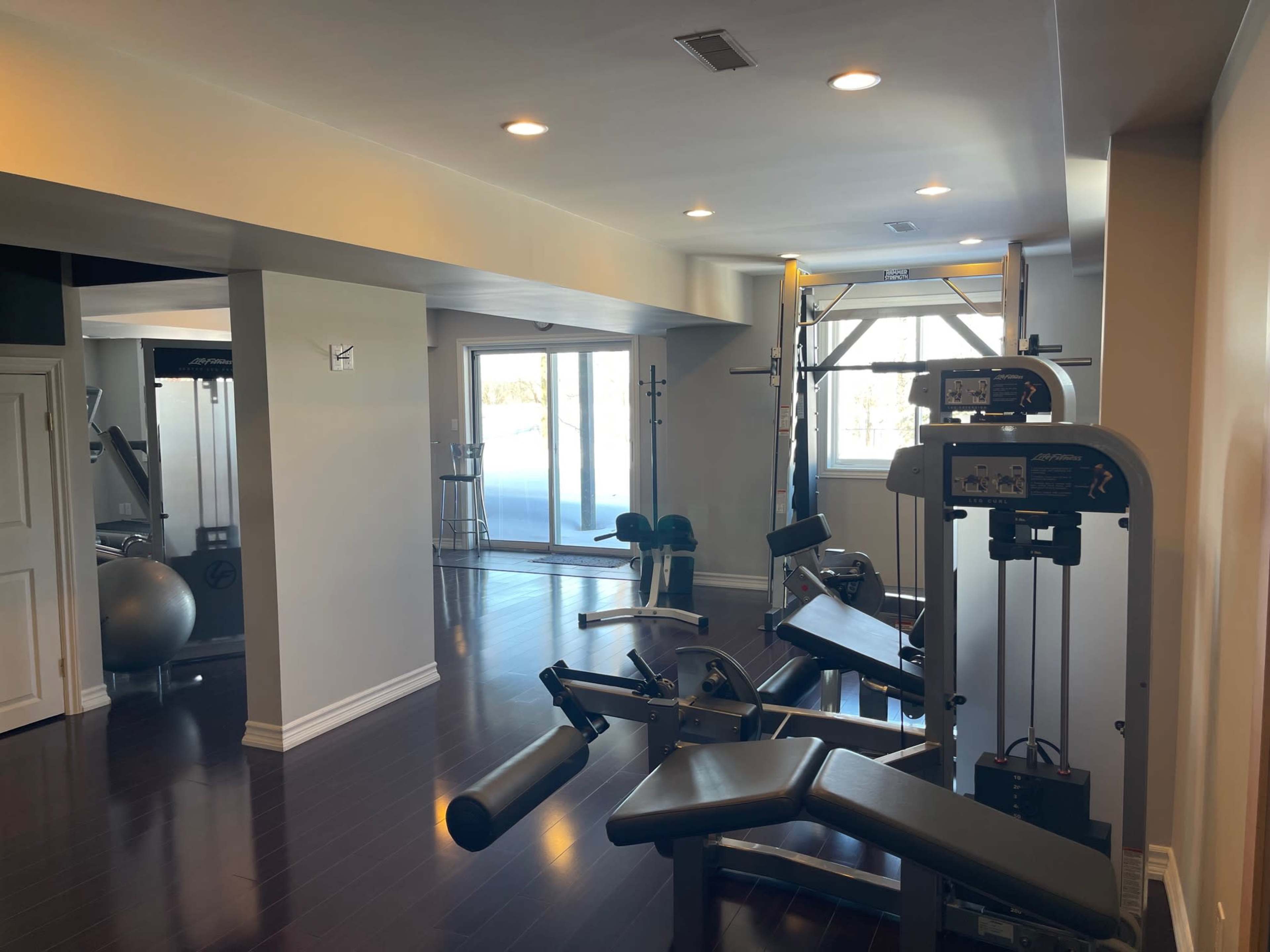 The image shows a home gym with various fitness equipment, including weight machines, benches, and a stability ball, in a well-lit room.