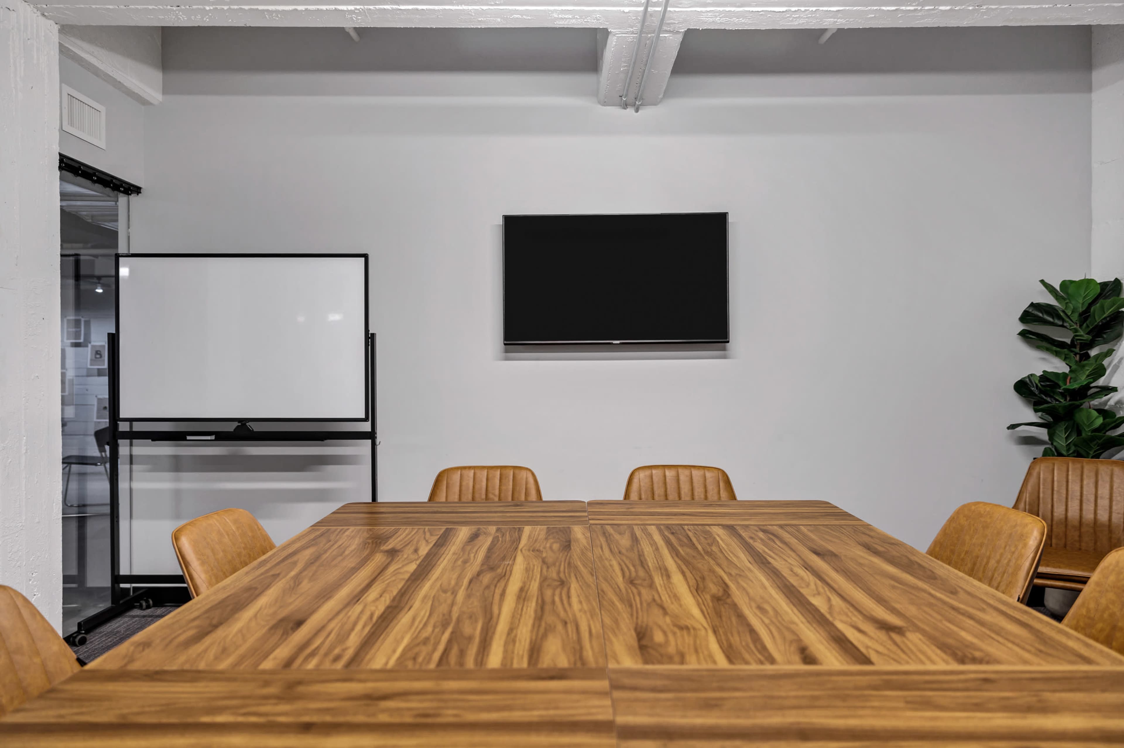 A modern conference room features a large wooden table surrounded by brown chairs, with a television mounted on the wall and a whiteboard on a stand nearby.