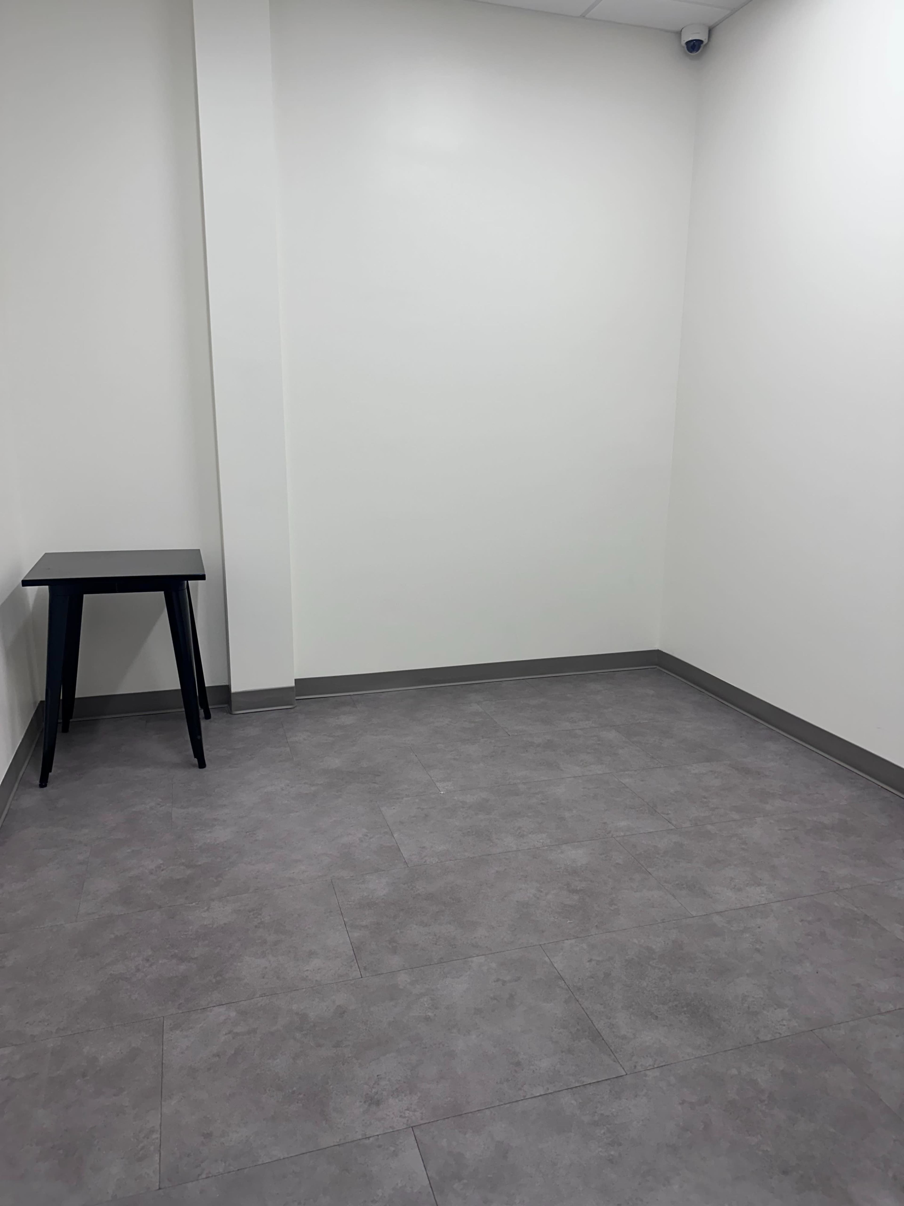 The image shows a sparsely furnished room with light gray walls, a plain gray floor, and a black table positioned in one corner.