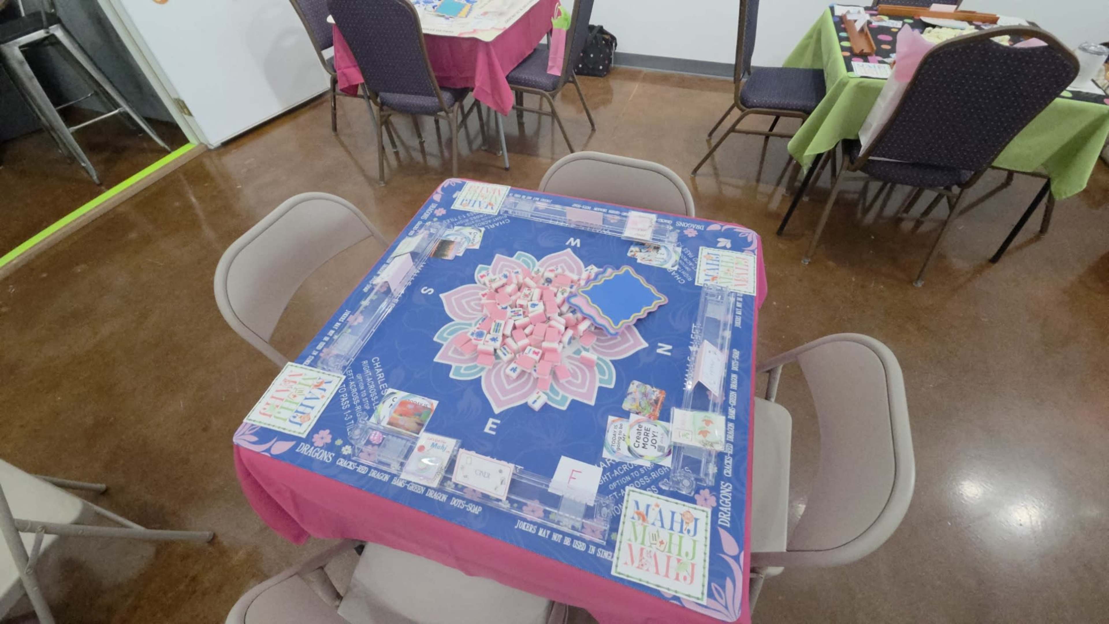 A game table is set up with a colorful board and various game pieces, surrounded by chairs in a brightly lit room with tables covered in cloths of different colors.