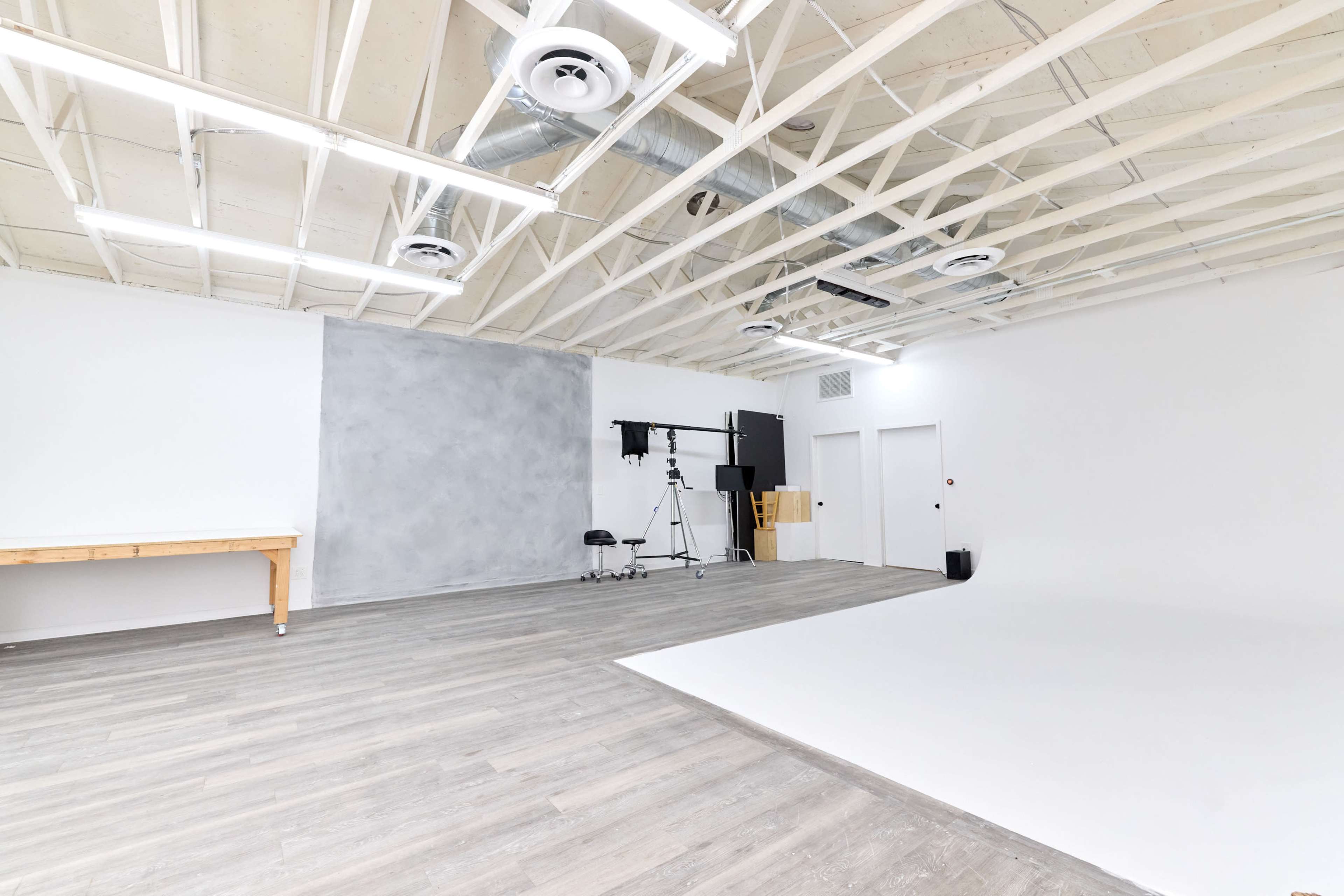 A spacious, well-lit photography studio featuring a gray backdrop, wooden table, and various equipment arranged in a minimalist layout.