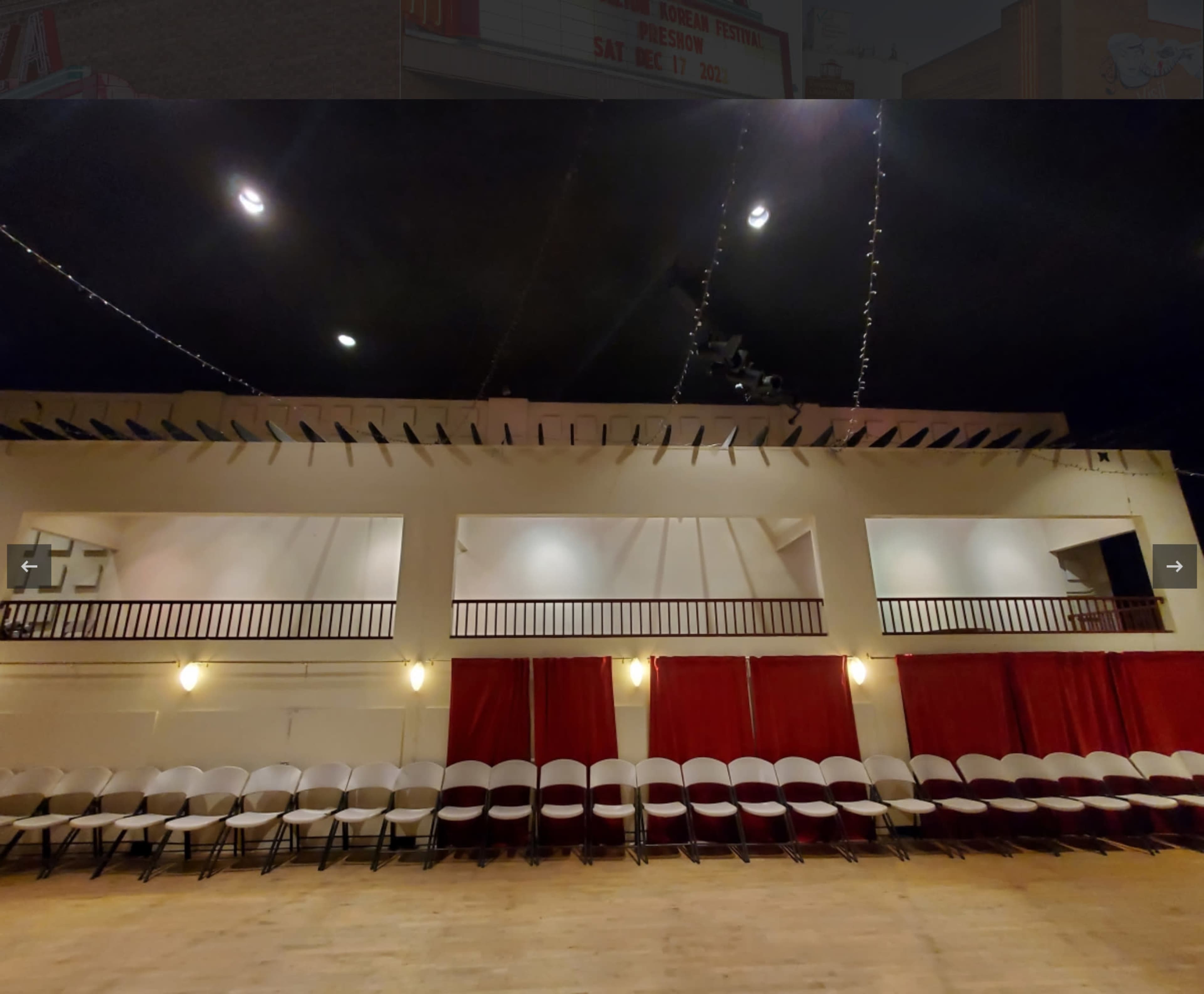 The image shows a spacious indoor venue with rows of white chairs set up facing a stage area, featuring red curtains and a balcony above.