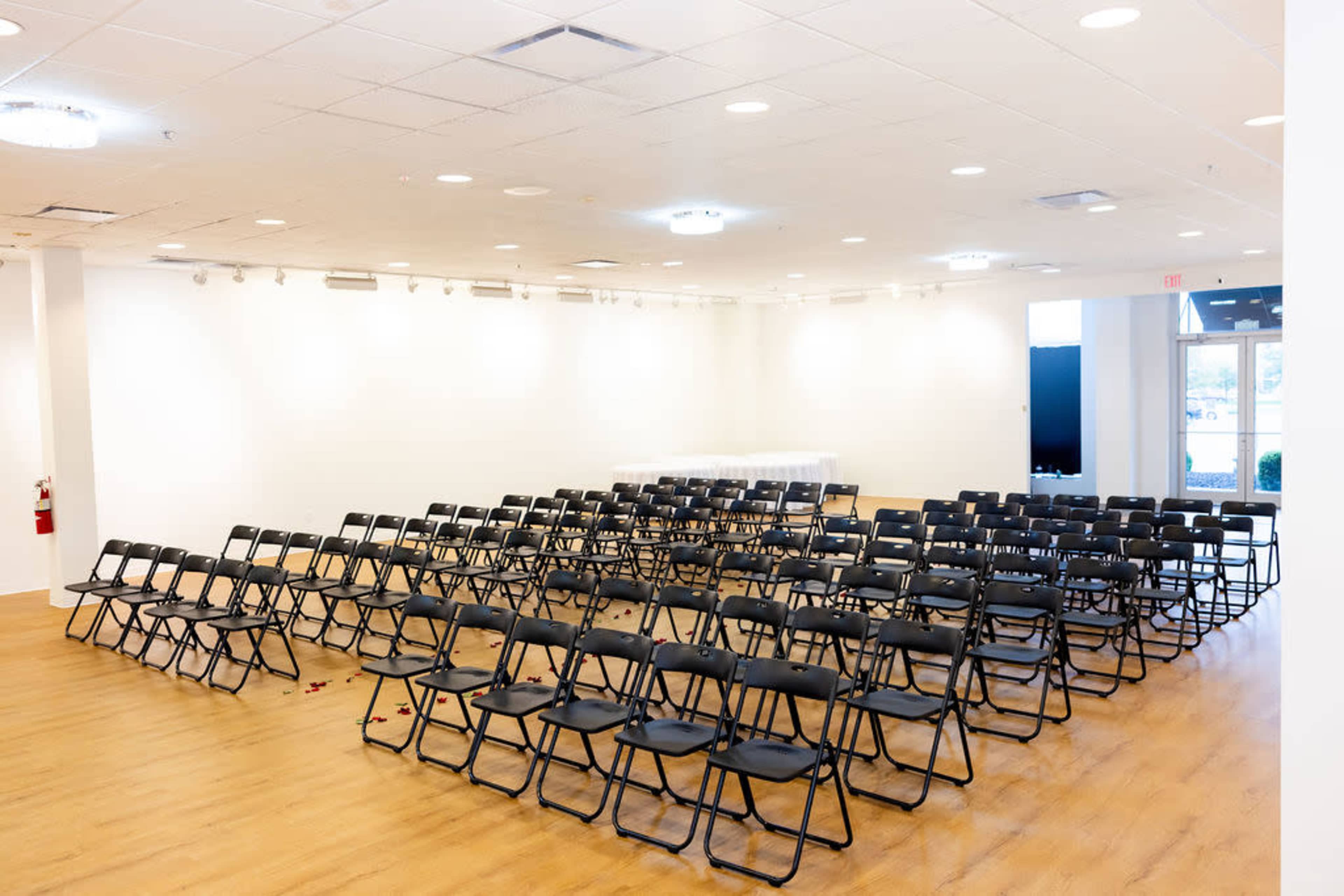 The image shows a spacious room filled with rows of black folding chairs arranged for an event.