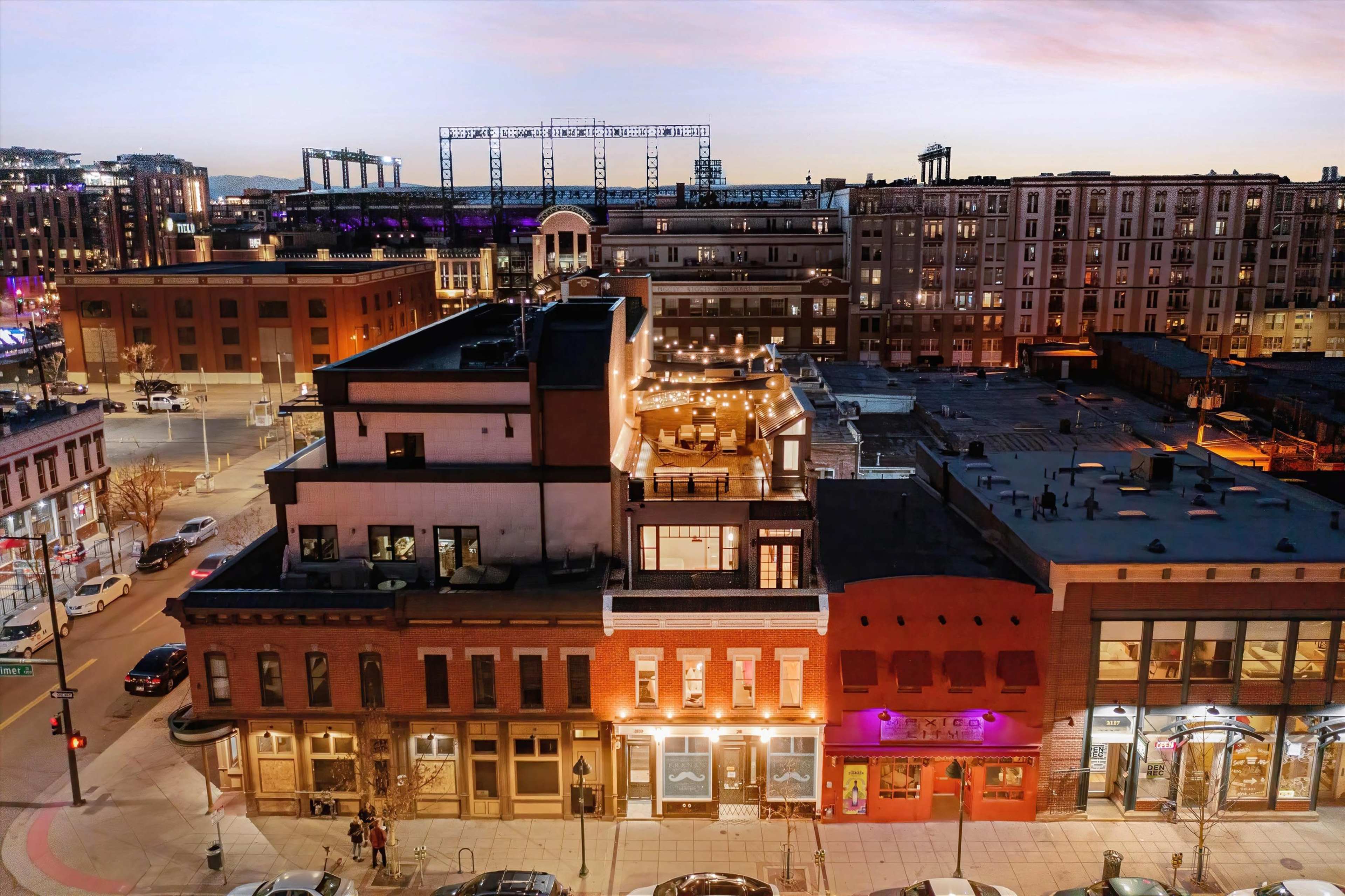 The image shows a rooftop restaurant atop a multi-story brick building in an urban area during twilight, with city lights visible in the background.