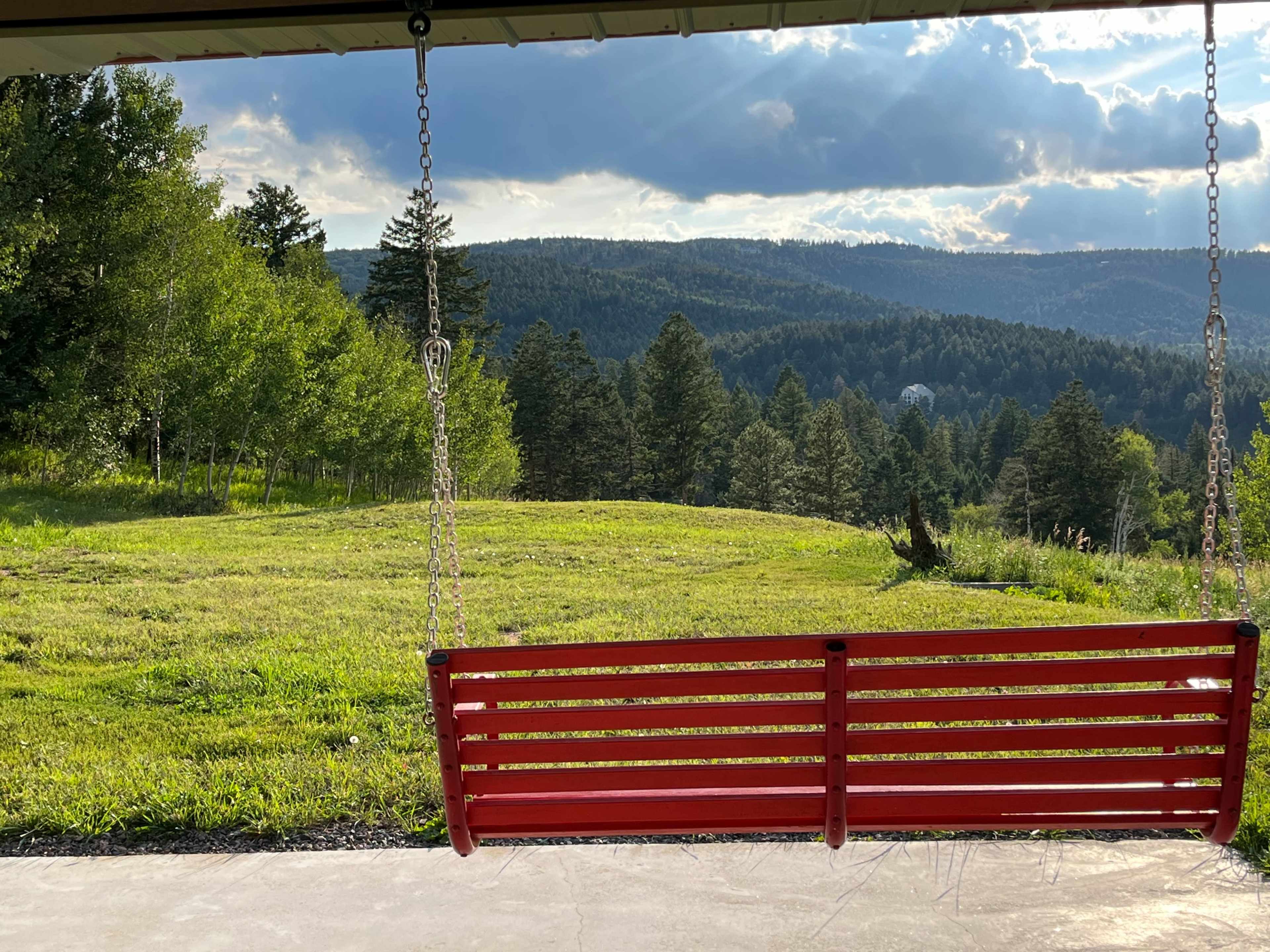 A red swing hangs from a covered porch overlooking a grassy field and wooded hills under a cloudy sky.
