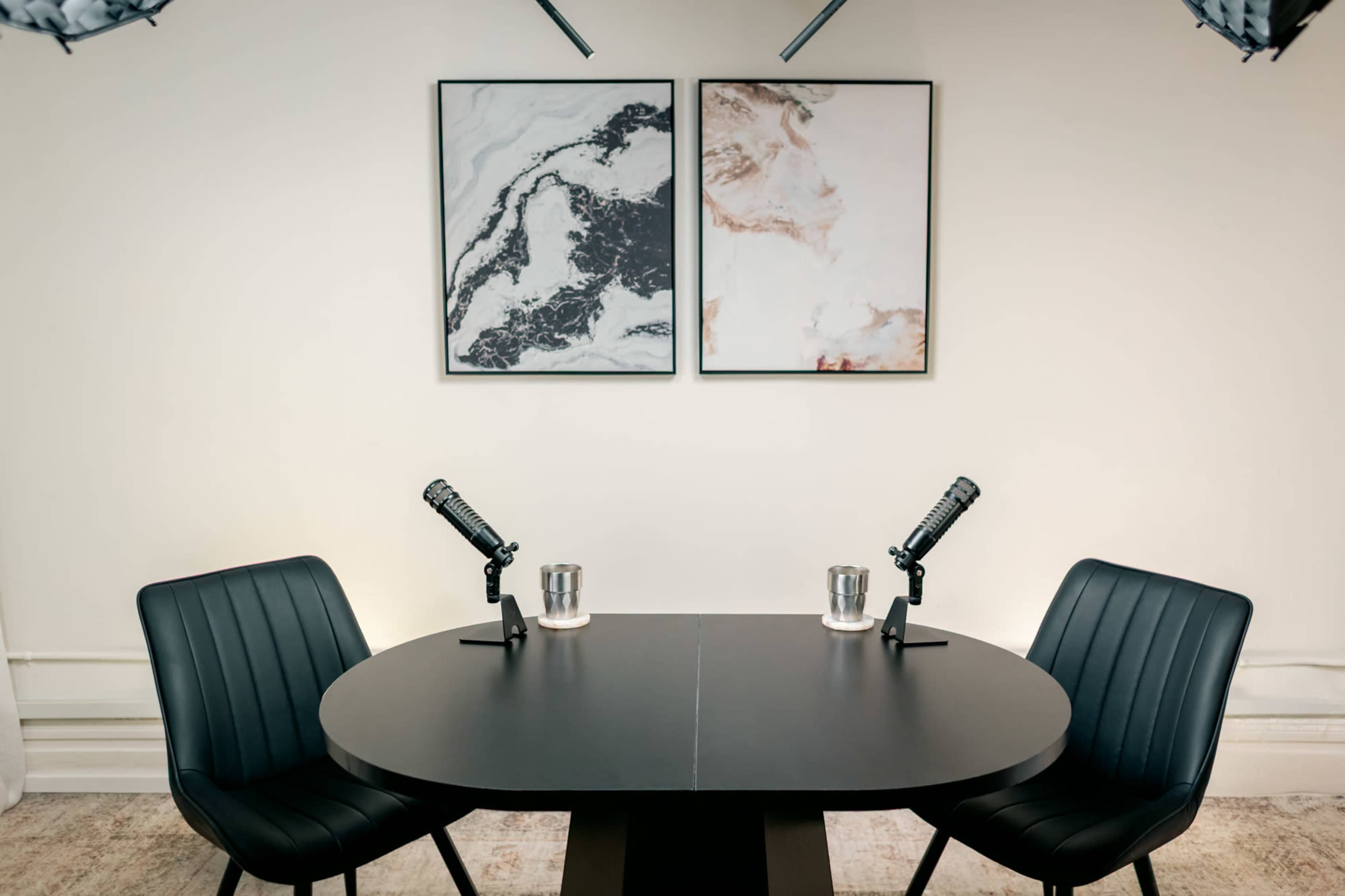 The image shows a minimalist podcast studio with two black chairs facing a round table, each equipped with a microphone and a glass of water, beneath two abstract wall art pieces.