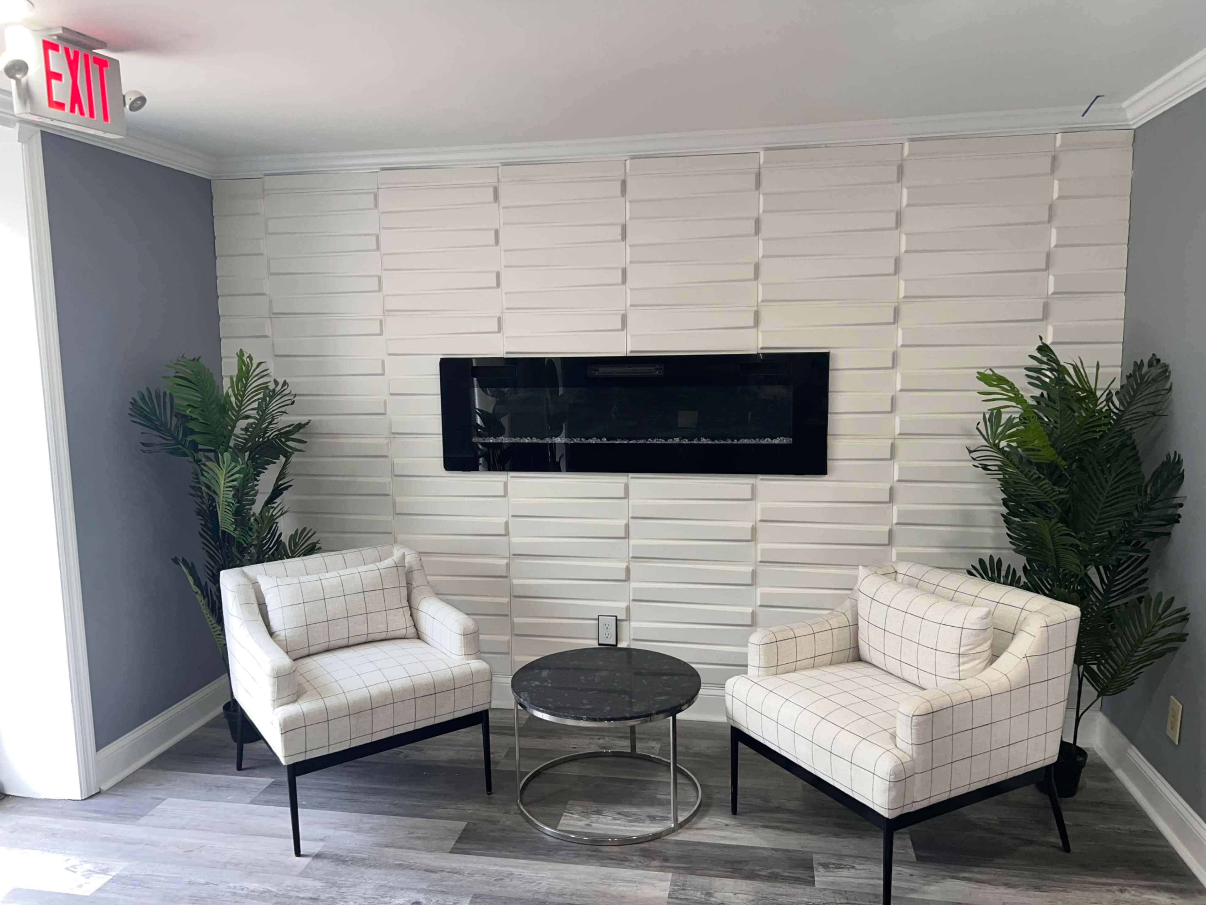 A minimalist seating area featuring two white checkered armchairs, a round black table, and a wall-mounted electric fireplace, flanked by green potted plants.
