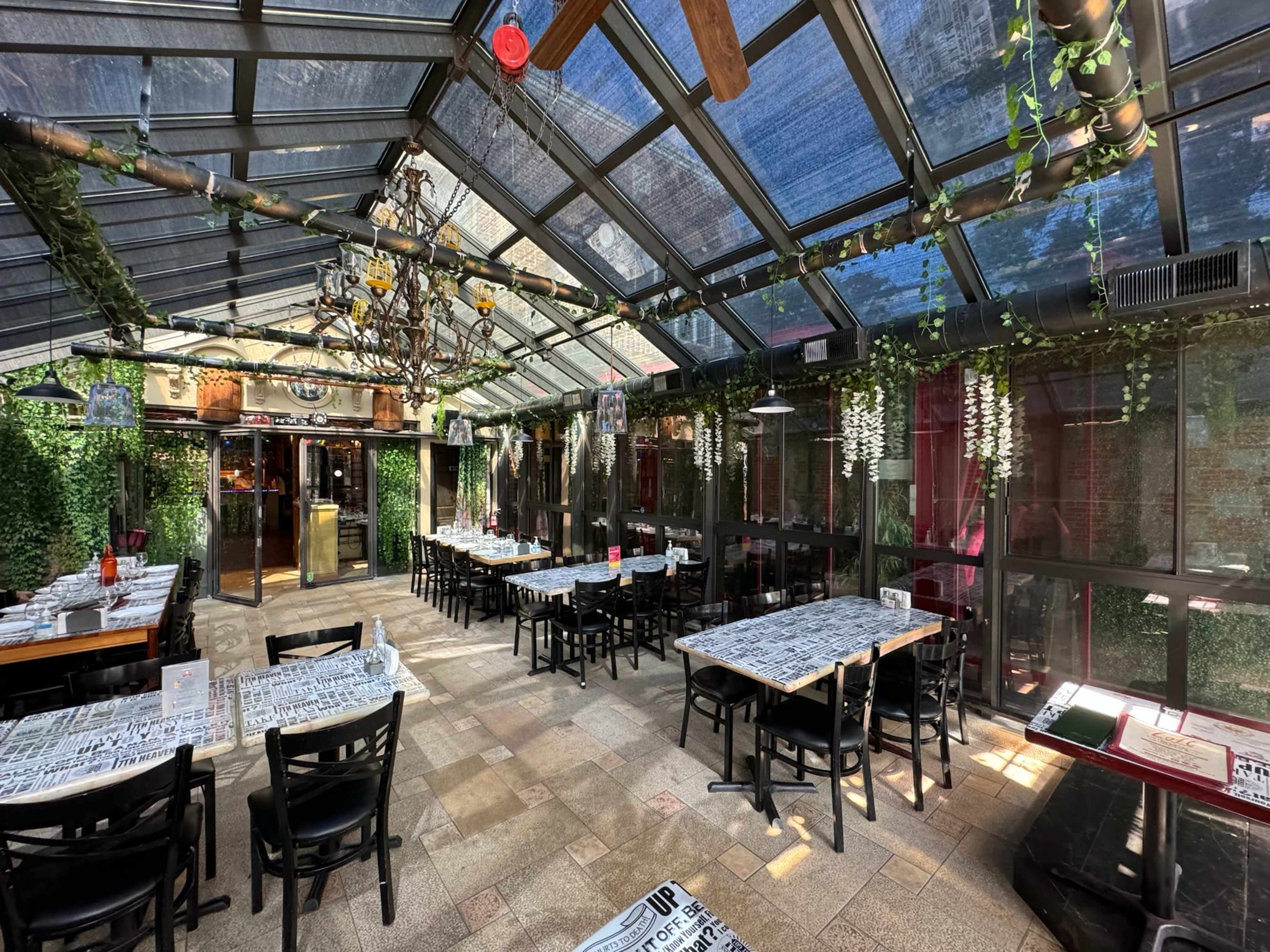 The image shows a bright, glass-enclosed dining area with wooden tables and black chairs, featuring greenery and decorative elements.