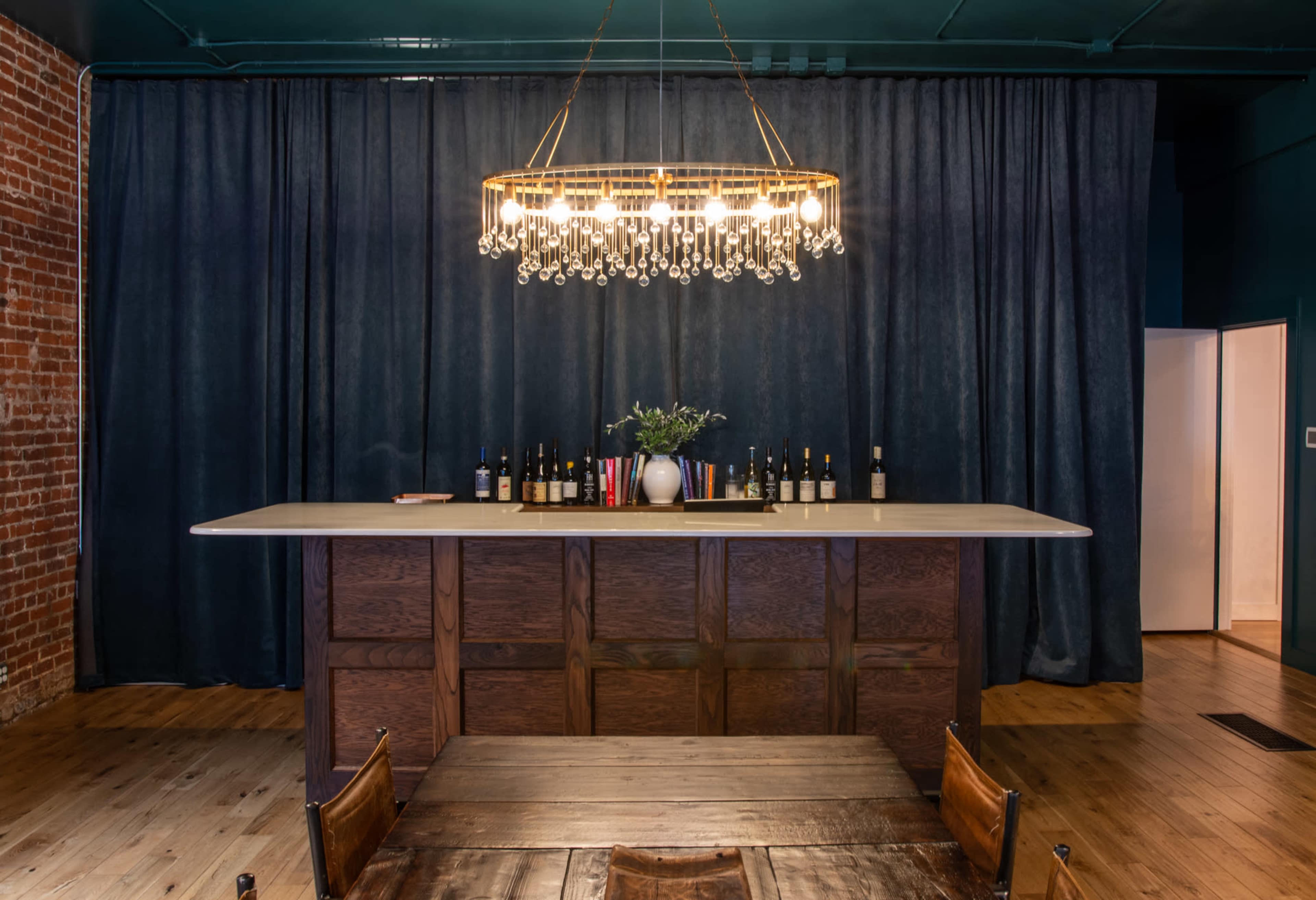 A modern bar area with a long, light-colored countertop, a chandelier above, and a backdrop of dark curtains and brick walls.